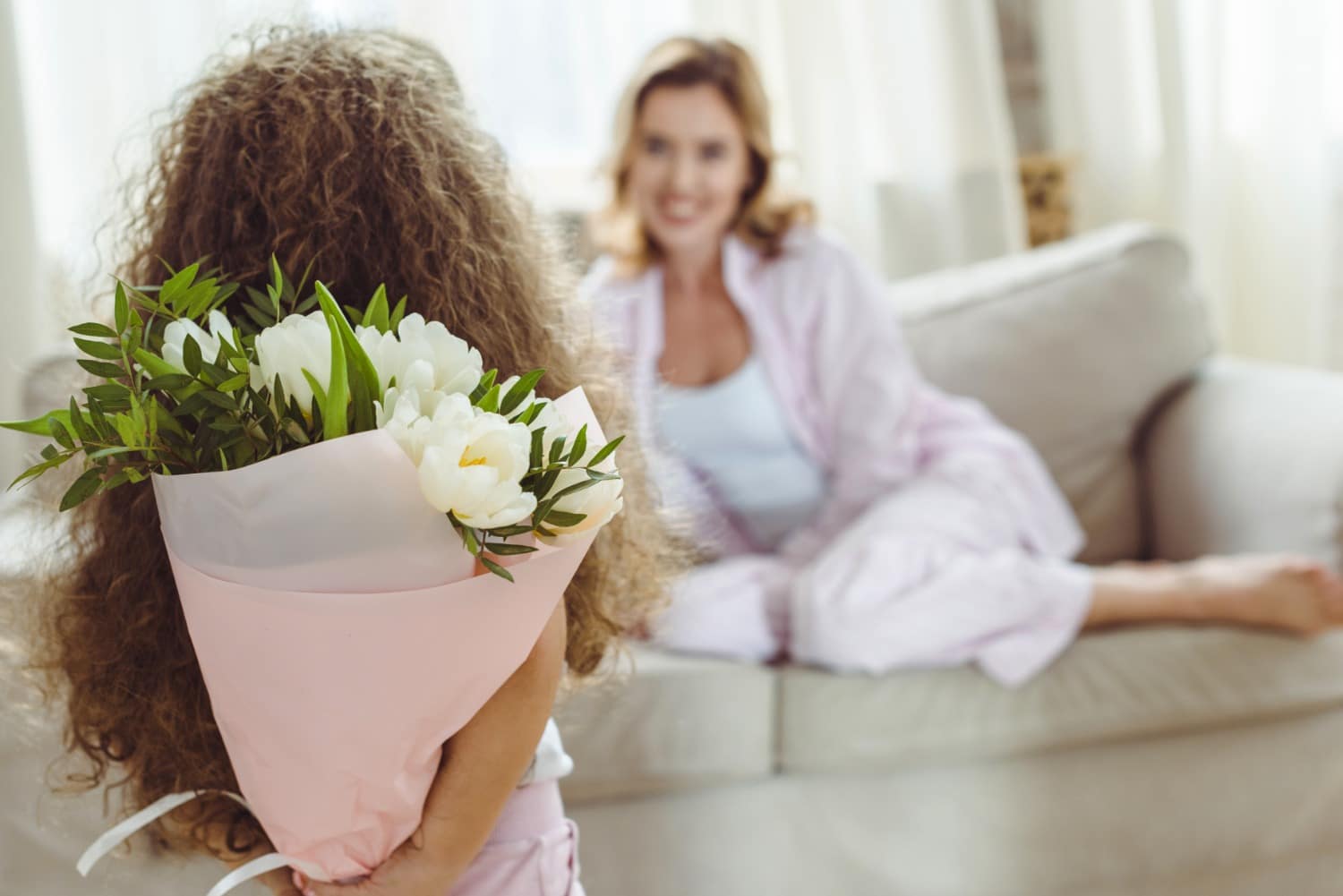 girl with flowers behind her back standing in foreground, white mom on couch in background