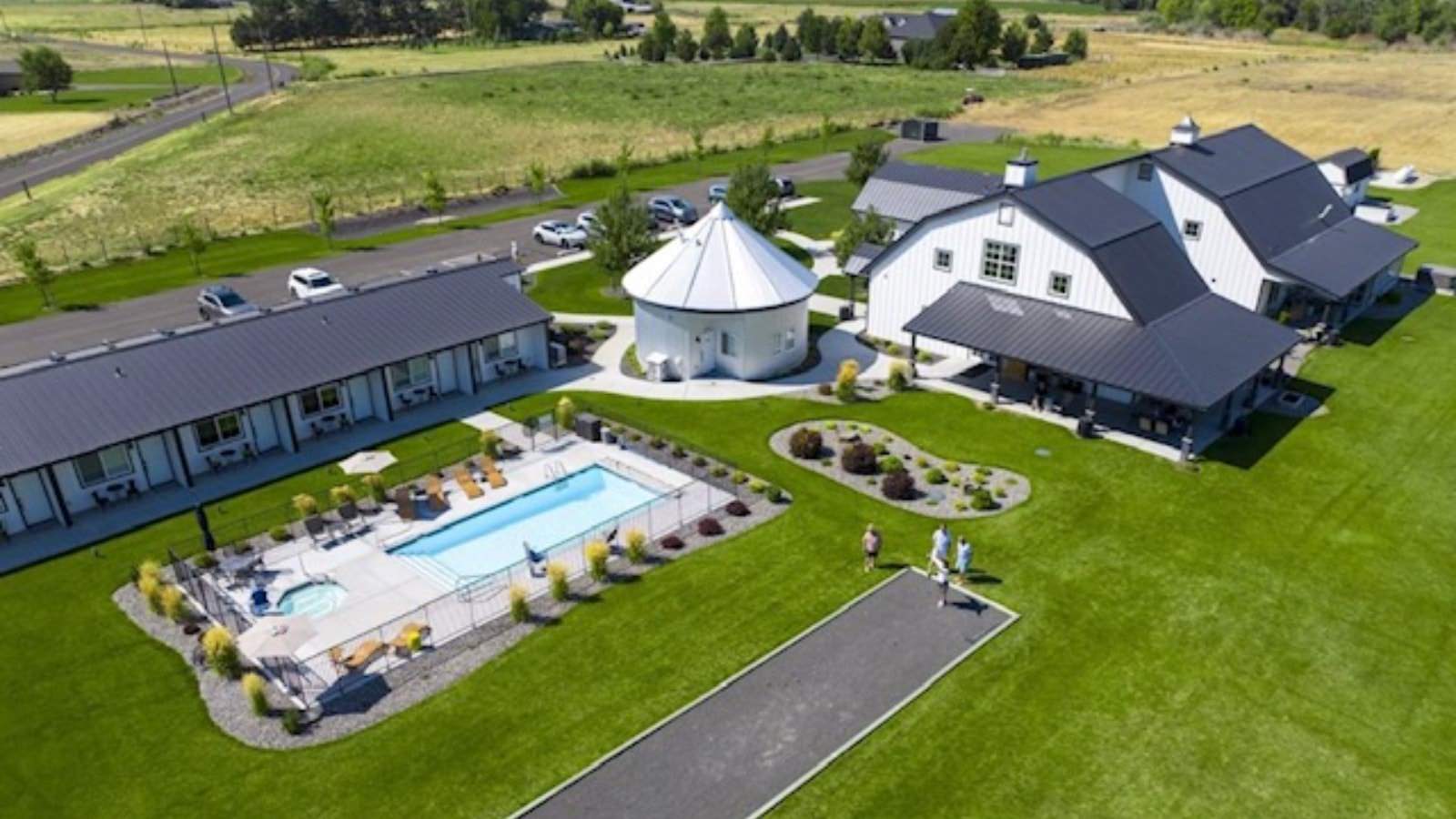 Aerial view of the property painted white with black trim, black roof, large covered porch, large green lawn, and fenced in swimming pool and hot tub