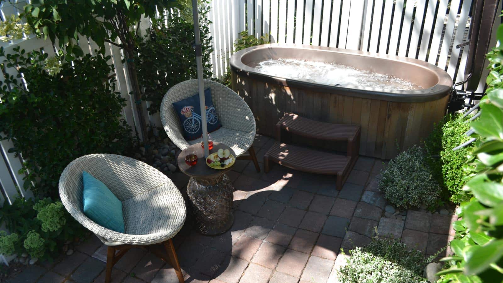 Brick patio with two-person hot tub and two wicker chairs surrounded by green vegetation