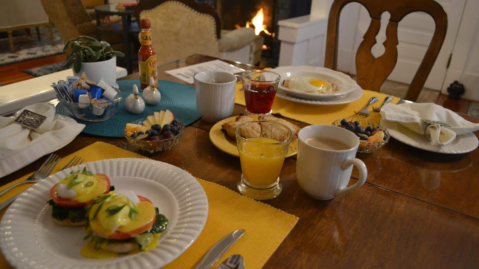 Close up view of breakfast dishes served on white plates on yellow placemats on wooden table with fireplace in the background