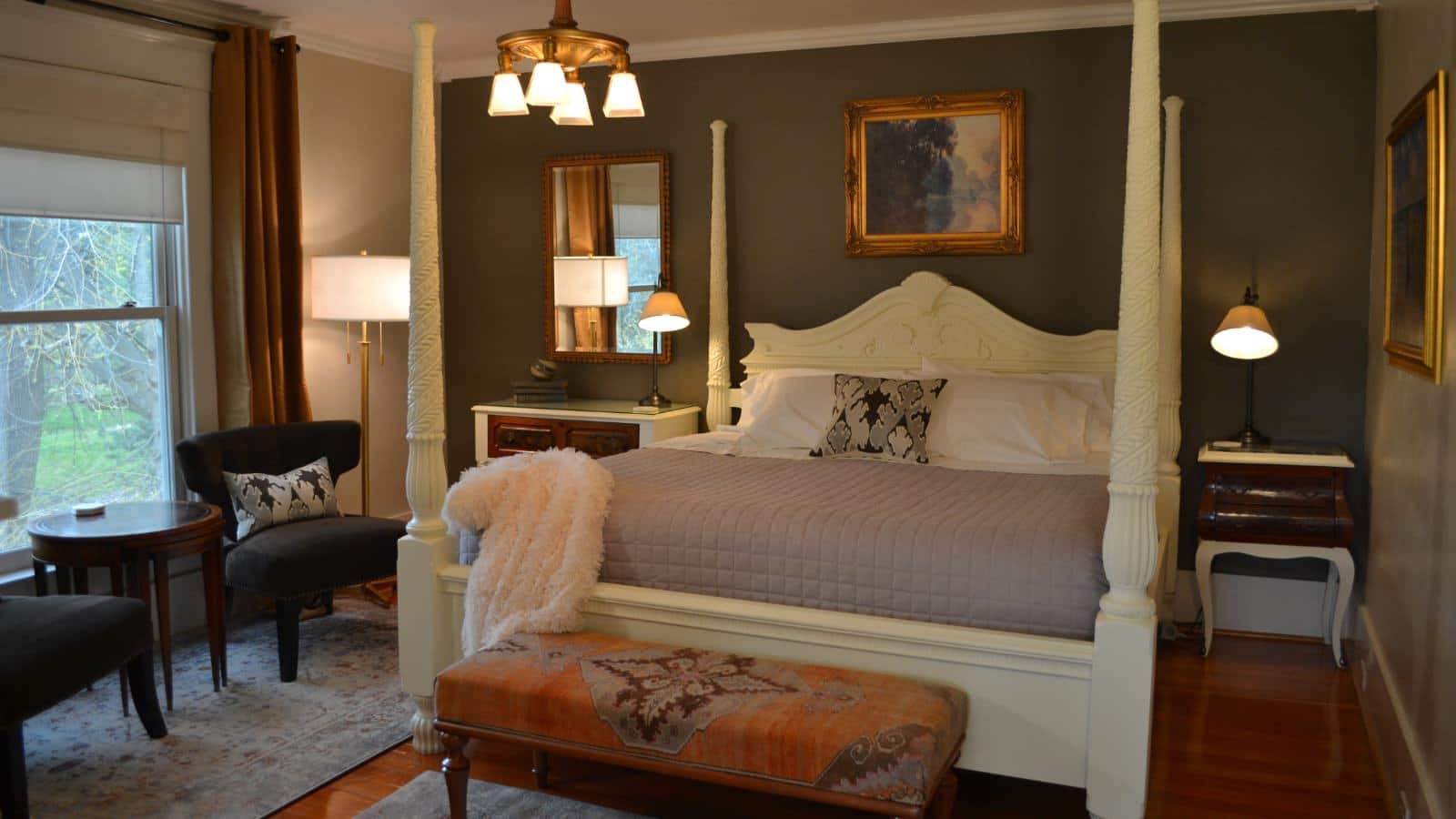 Bedroom with dark gray accent wall, hardwood flooring, white four-poster bed, white bedding, sitting area, and large window