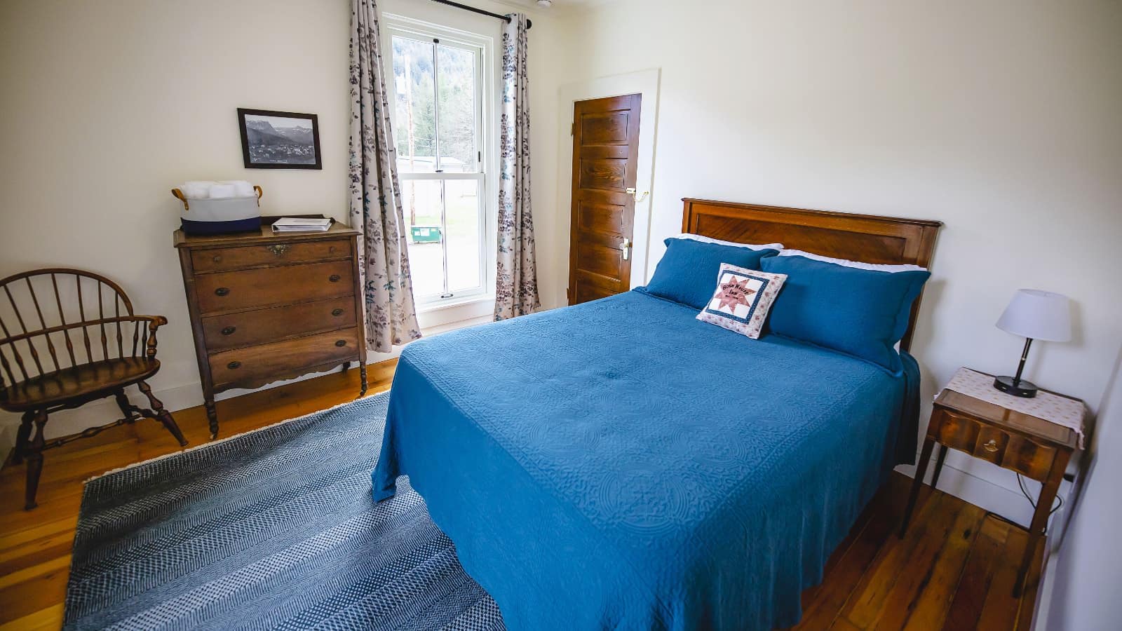 Bedroom with white walls, hardwood flooring, wooden bed, blue bedding, dark wooden dresser, and large window