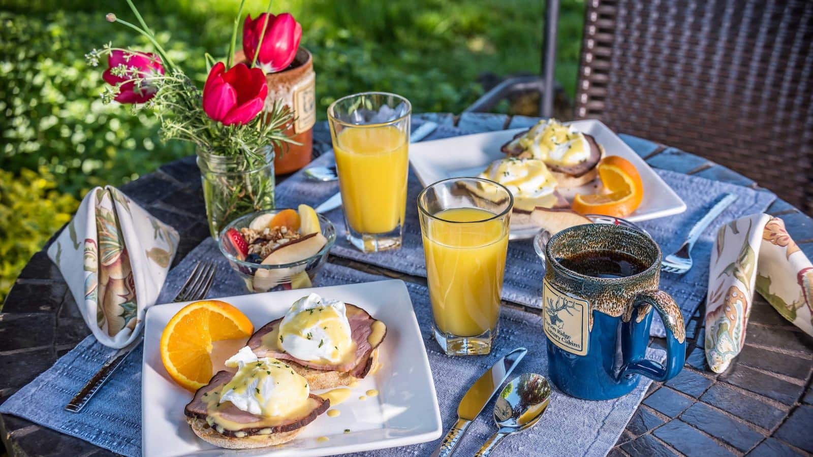 White square plates with breakfast dishes sitting on light blue placemats with orange juice and coffee all on dark brown mosaic stone table