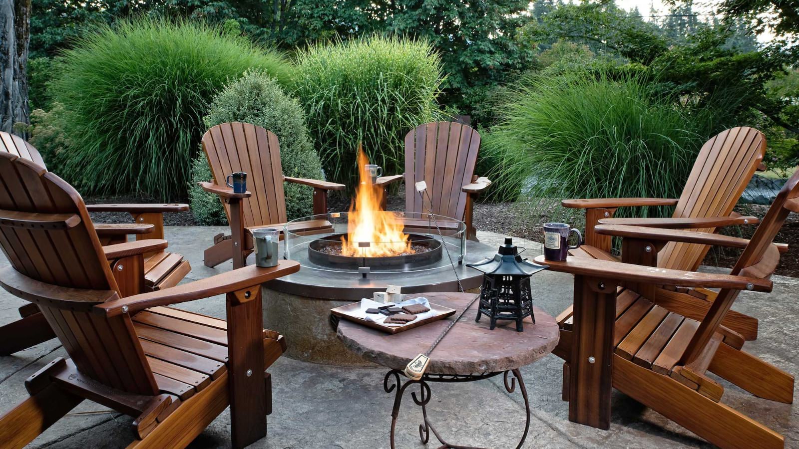 Multiple dark wooden Adirondack chairs around a fire pit with large green grasses in the background