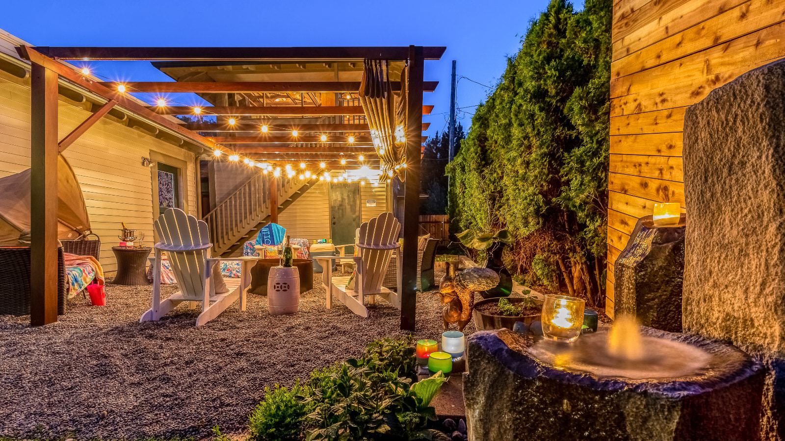 Back gravel patio with firepit and white Adirondack chairs under pergola with string lights next to a water fountain and green plants