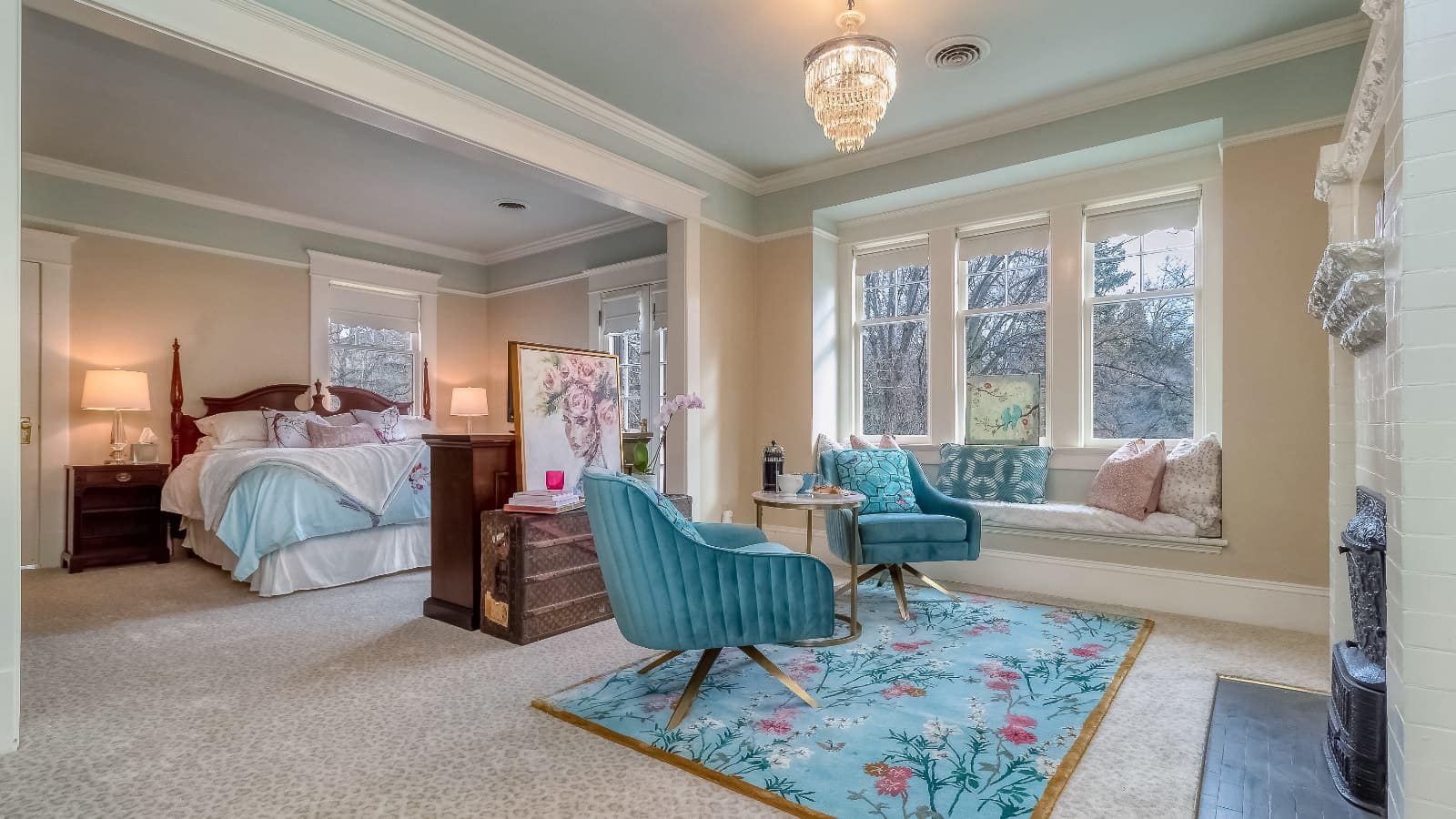 Bedroom suite with light peach walls, white trim, carpeting, dark wooden bed, white bedding, turquoise upholstered chairs, fireplace, and large windows