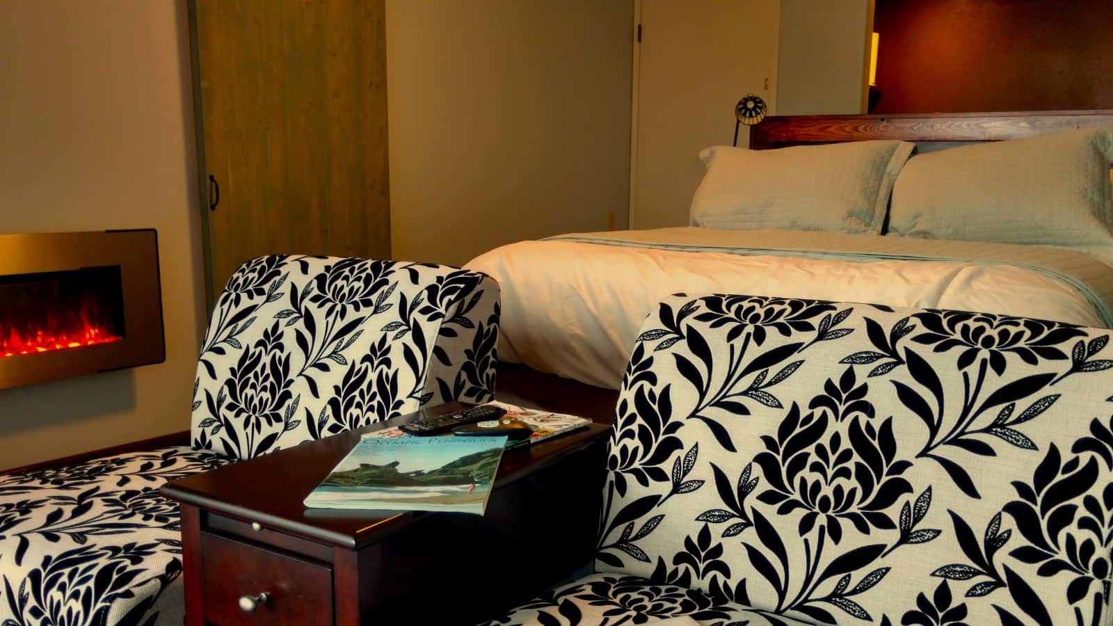 Bedroom with white walls, wooden bed, light colored bedding, fireplace, and sitting area with black and white floral patterned upholstered chairs