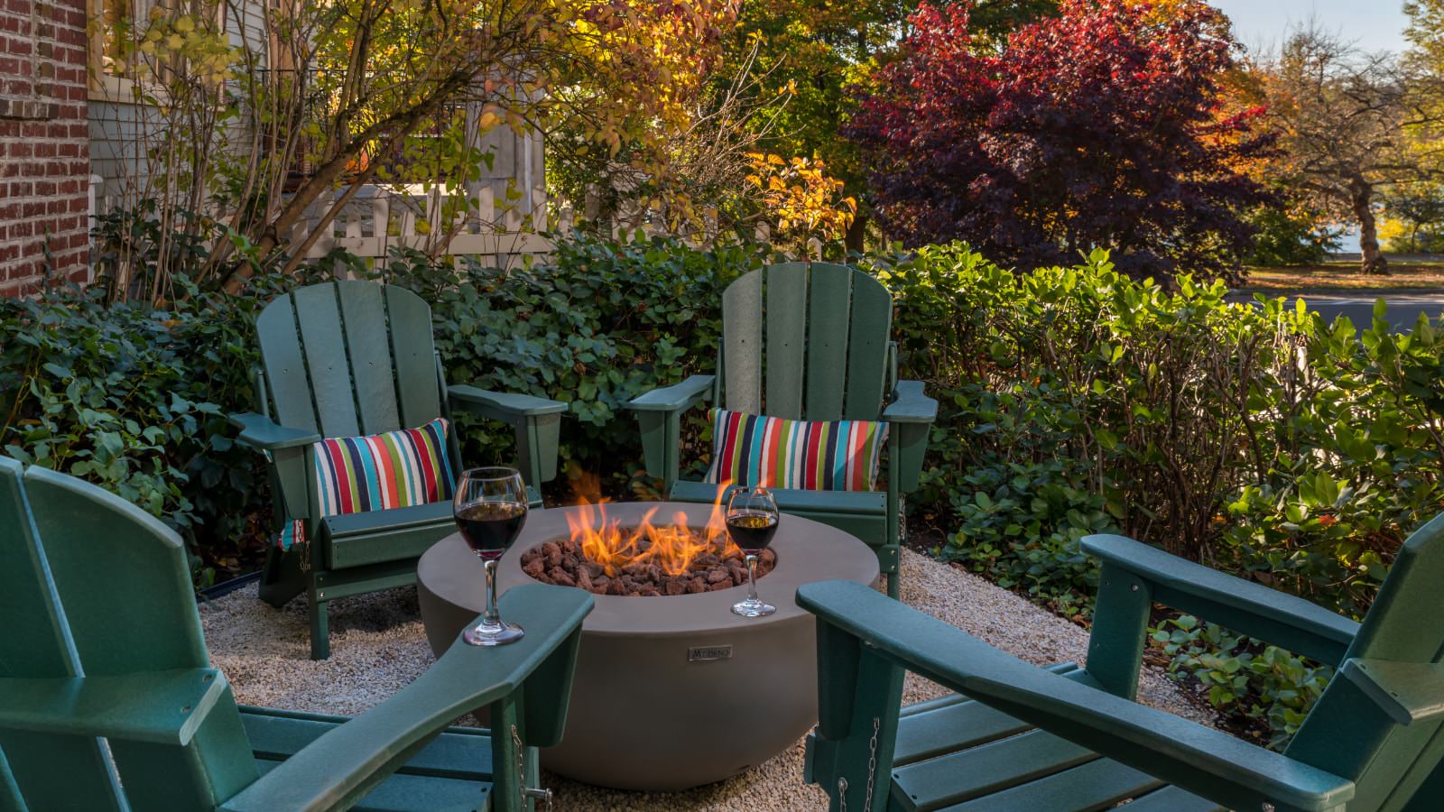 Gravel patio with light brown circular fire pit surrounded by four dark green Adirondack chairs