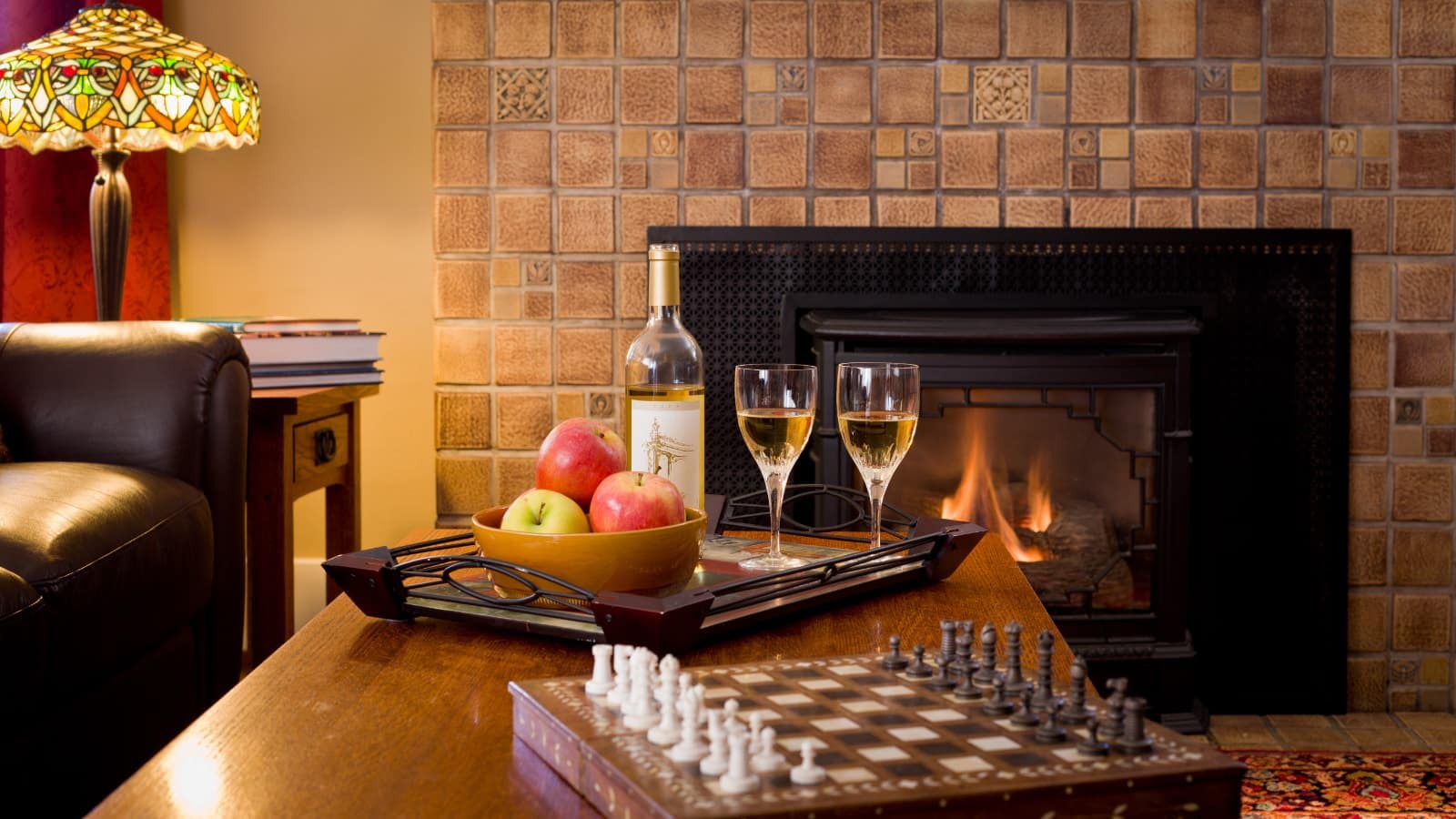 Close up view of wooden coffee table with chess board and serving tray with bowl of fruit and white wine next to fireplace