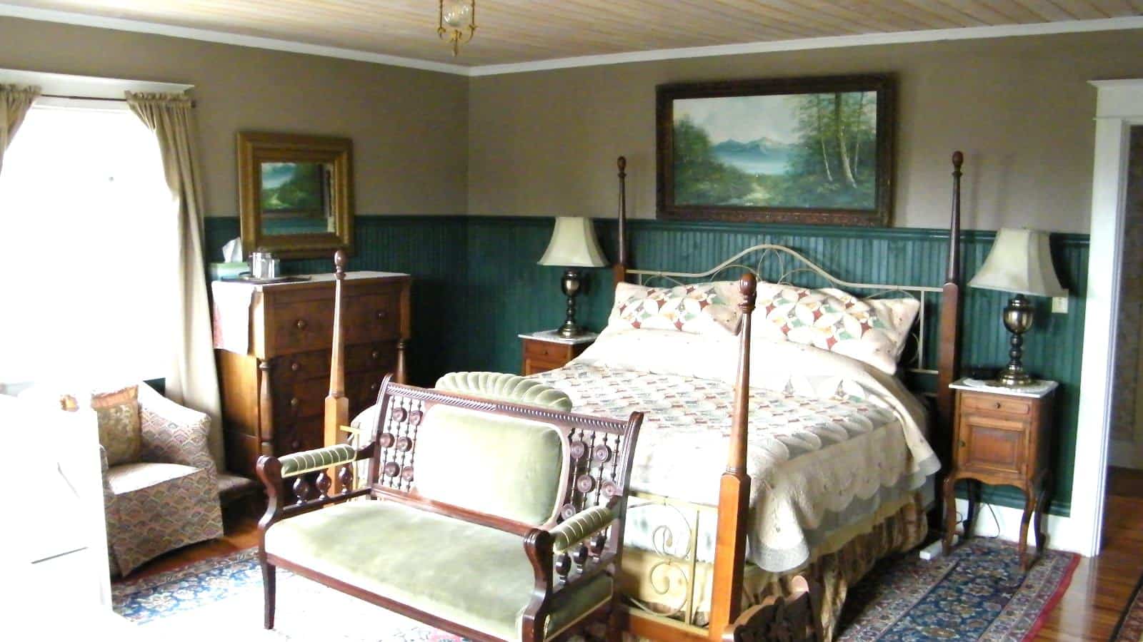 Bedroom with green wainscotting and light taupe walls, hardwood flooring, wooden four-poster bed, multicolored bedding, and wooden dresser