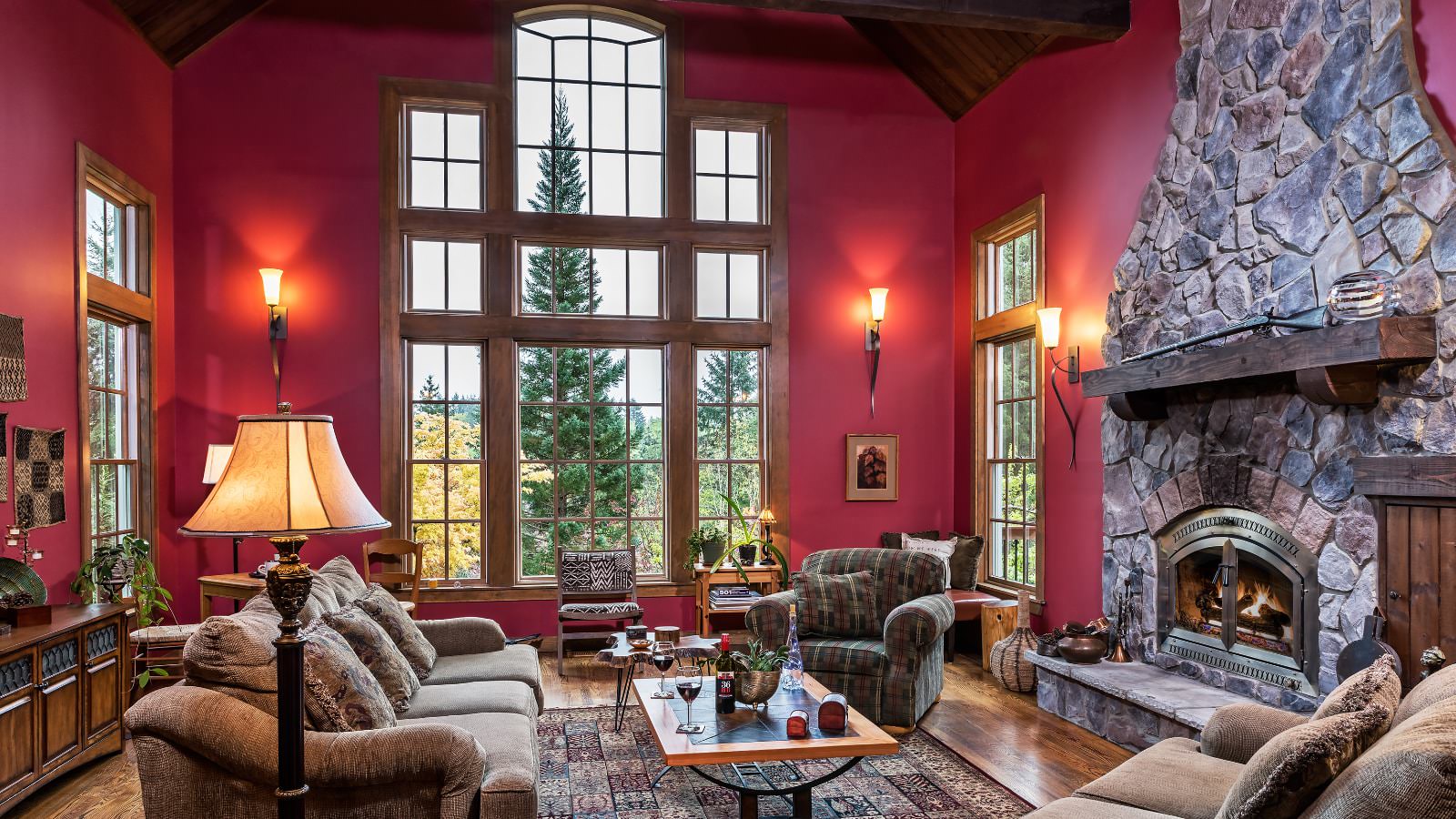 Large great room with dark pink walls, hardwood flooring, large stone fireplace, oversized couch and chairs, and large window with views to the outside