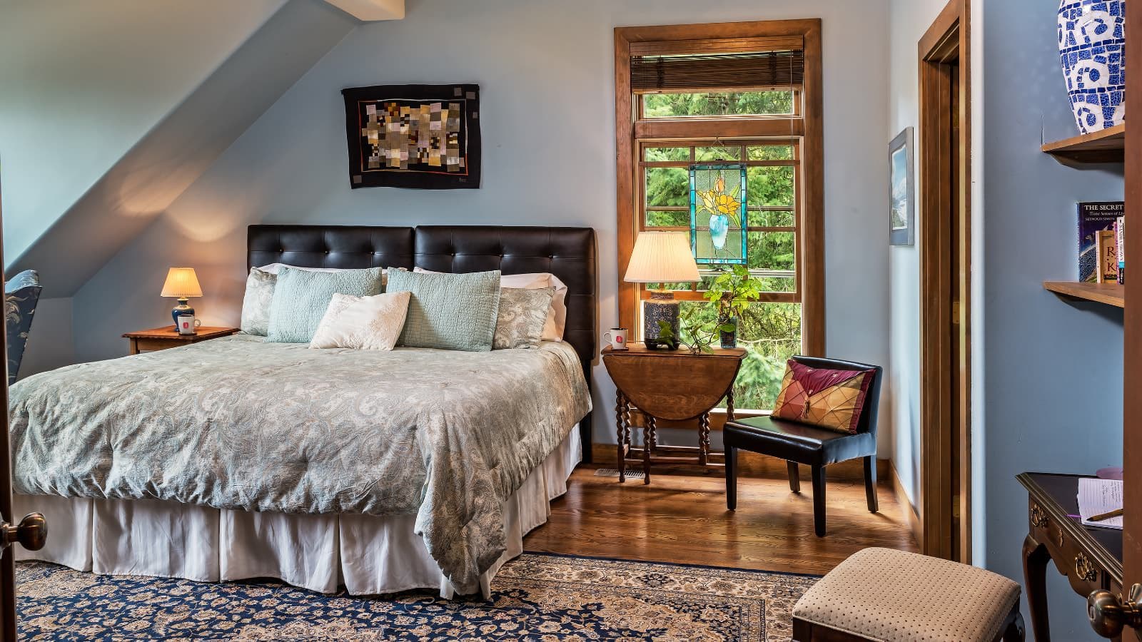 Bedroom with light blue walls, hardwood flooring, black leather headboard, light paisley bedding, and large window with view of the outside