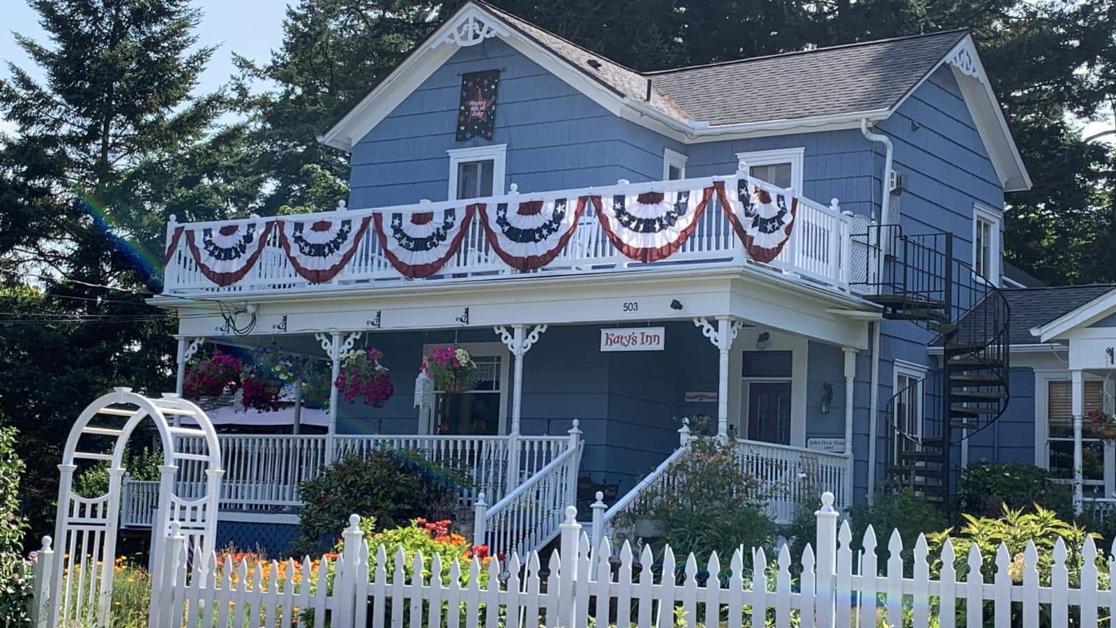 Exterior view of property painted light blue with white trim, white railed front porch and second-level balcony, and surrounded by green bushes, flowers, and trees