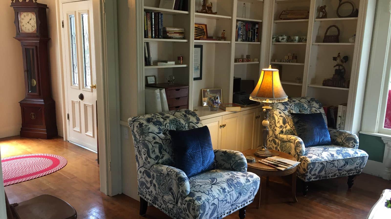 Living room with white walls, hardwood flooring, built-in shelving and cabinets, sitting area, and grandfather clock in foyer