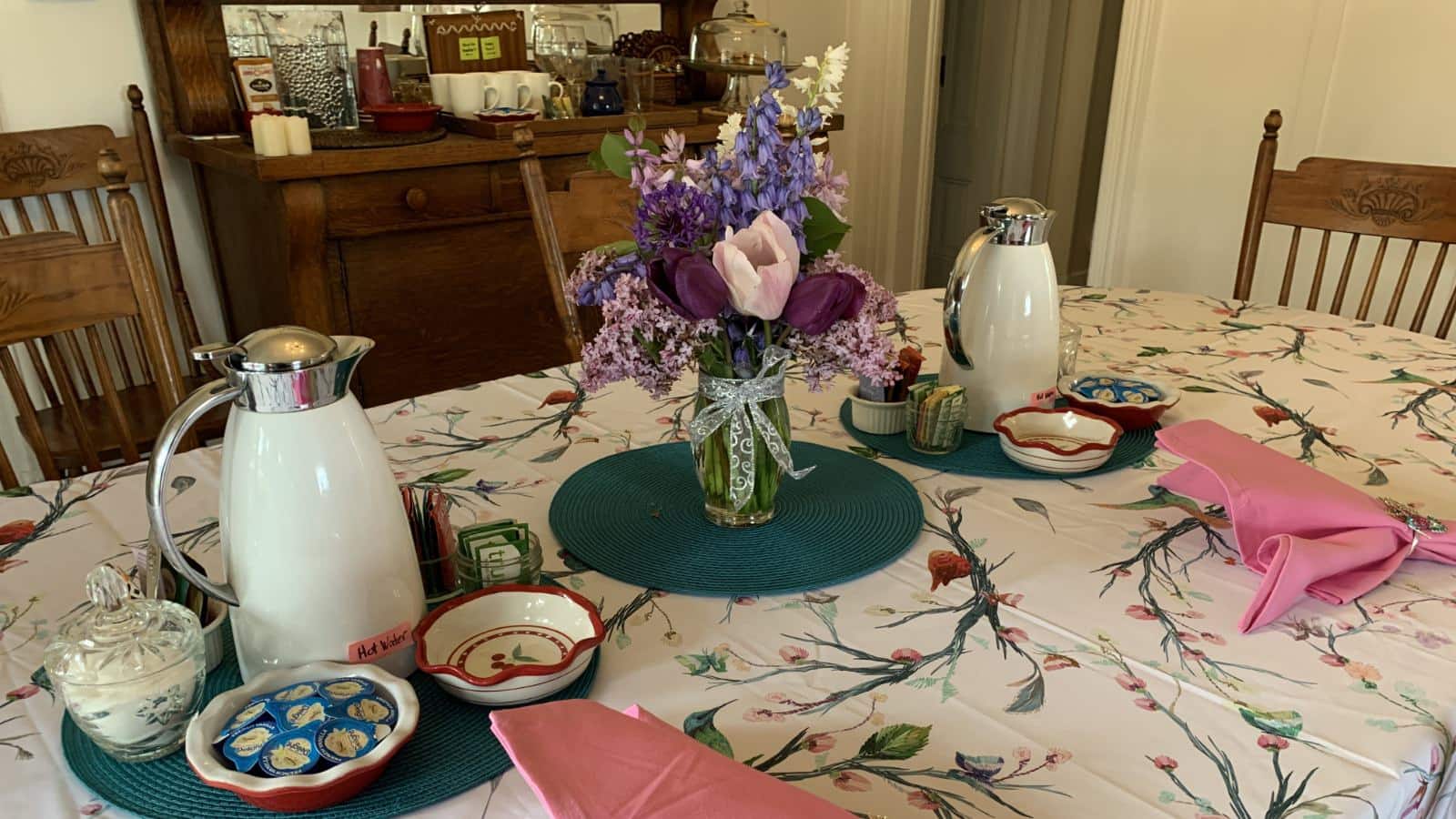 Close up of dining table with colorful tablecloth, pink cloth napkins, vase with purple flowers, white thermal pitchers with hot water, and dishes with tea and sugar packets