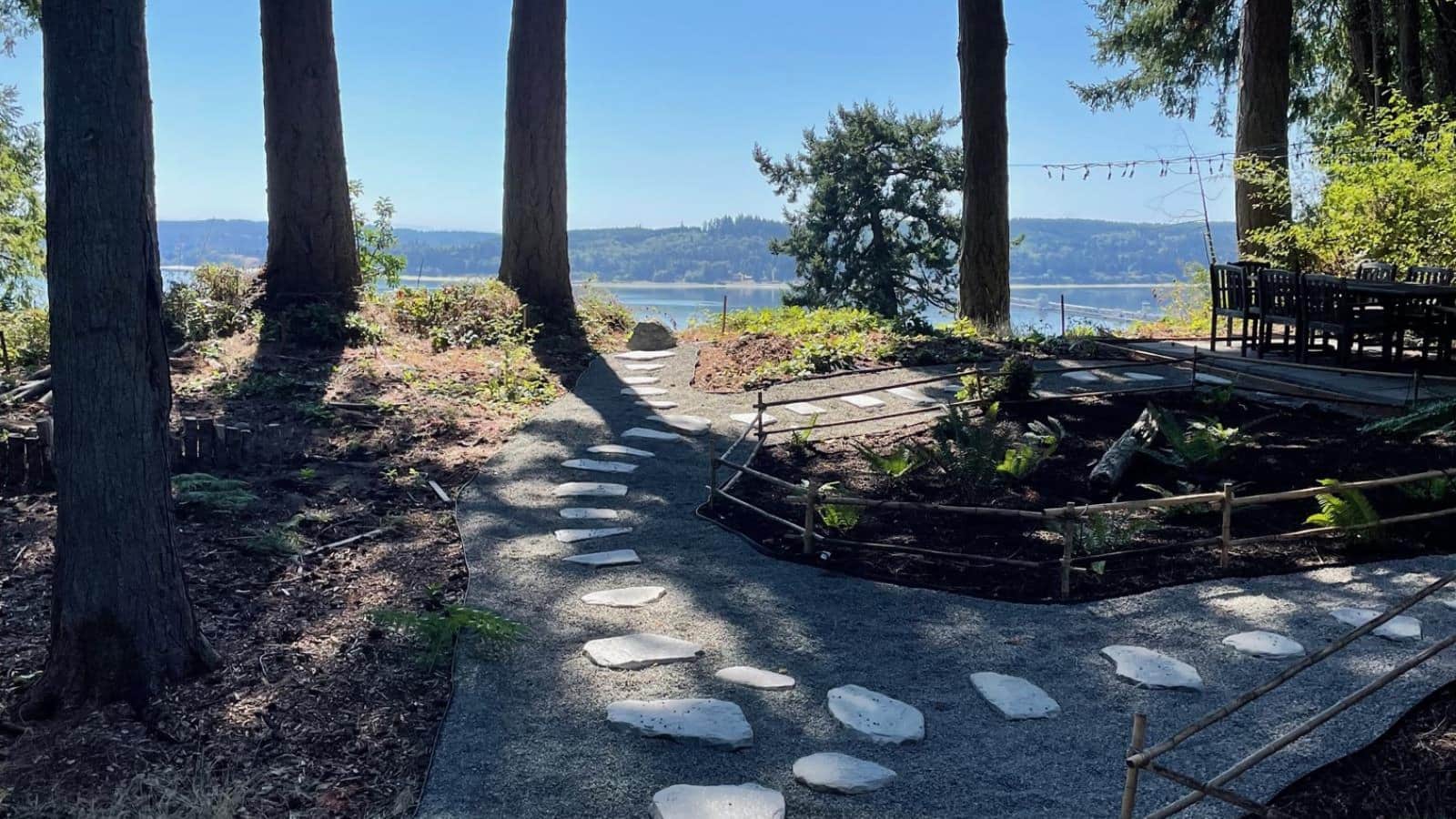 Gravel path shaded by large trees with stepping stones surrounding a flower garden with views of the water in the background