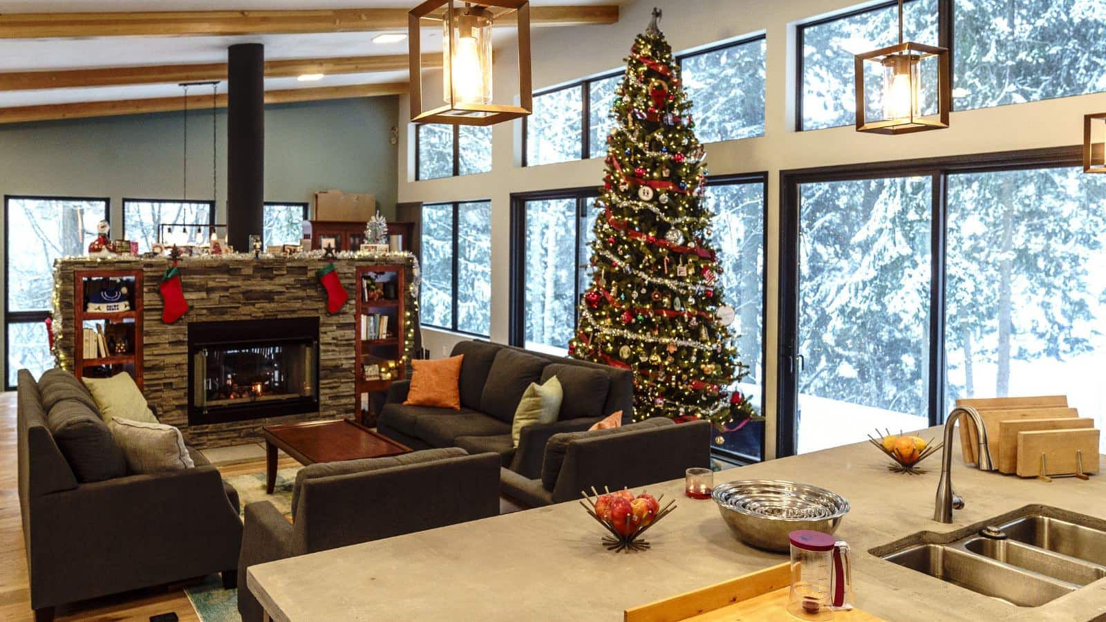 Open room with kitchen and living room with dark gray upholstered sectional, stone fireplace, large Christmas tree, and many large windows