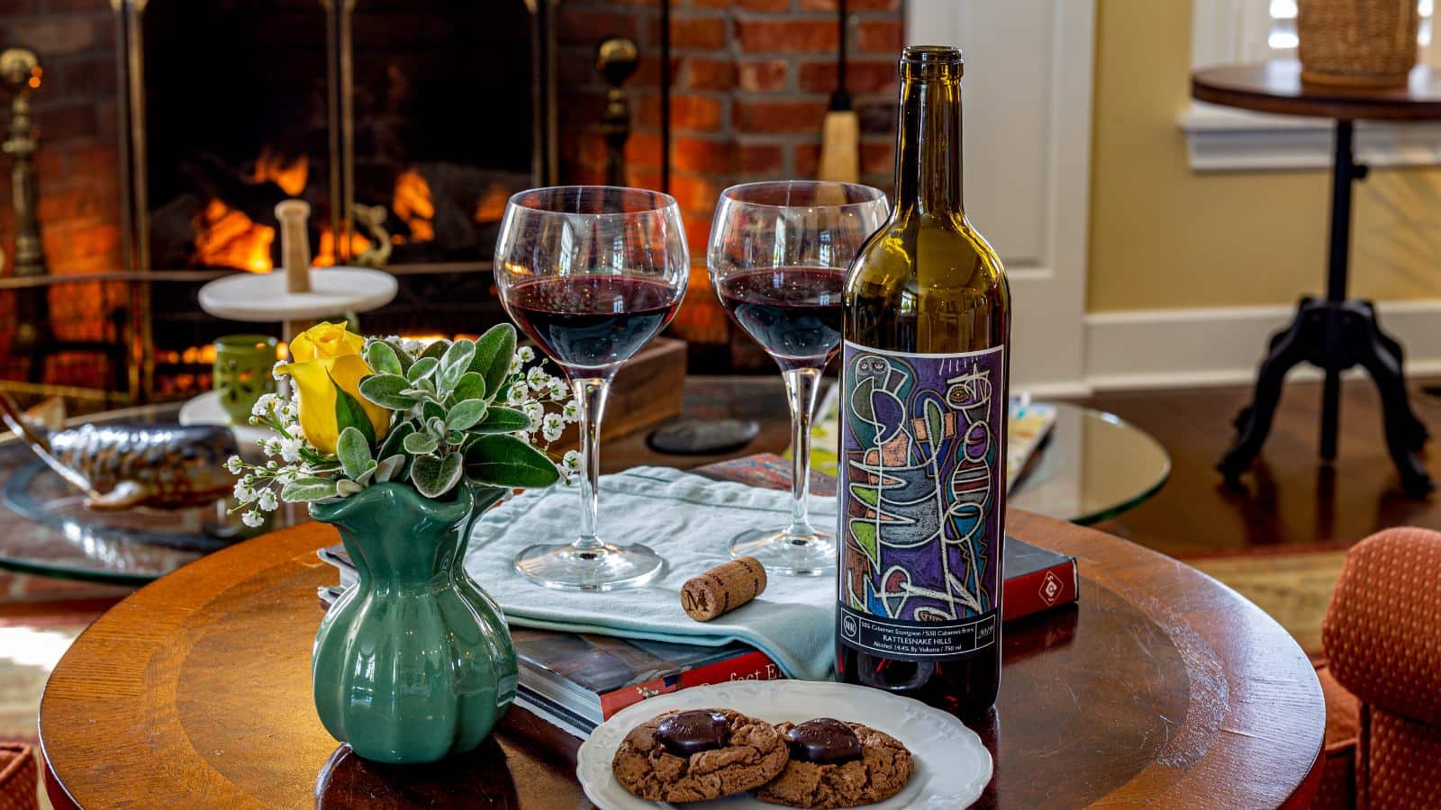 Close up view of small round wooden table with a small white plate of cookie, sage green vase with flowers, a book, and two glasses filled with red wine