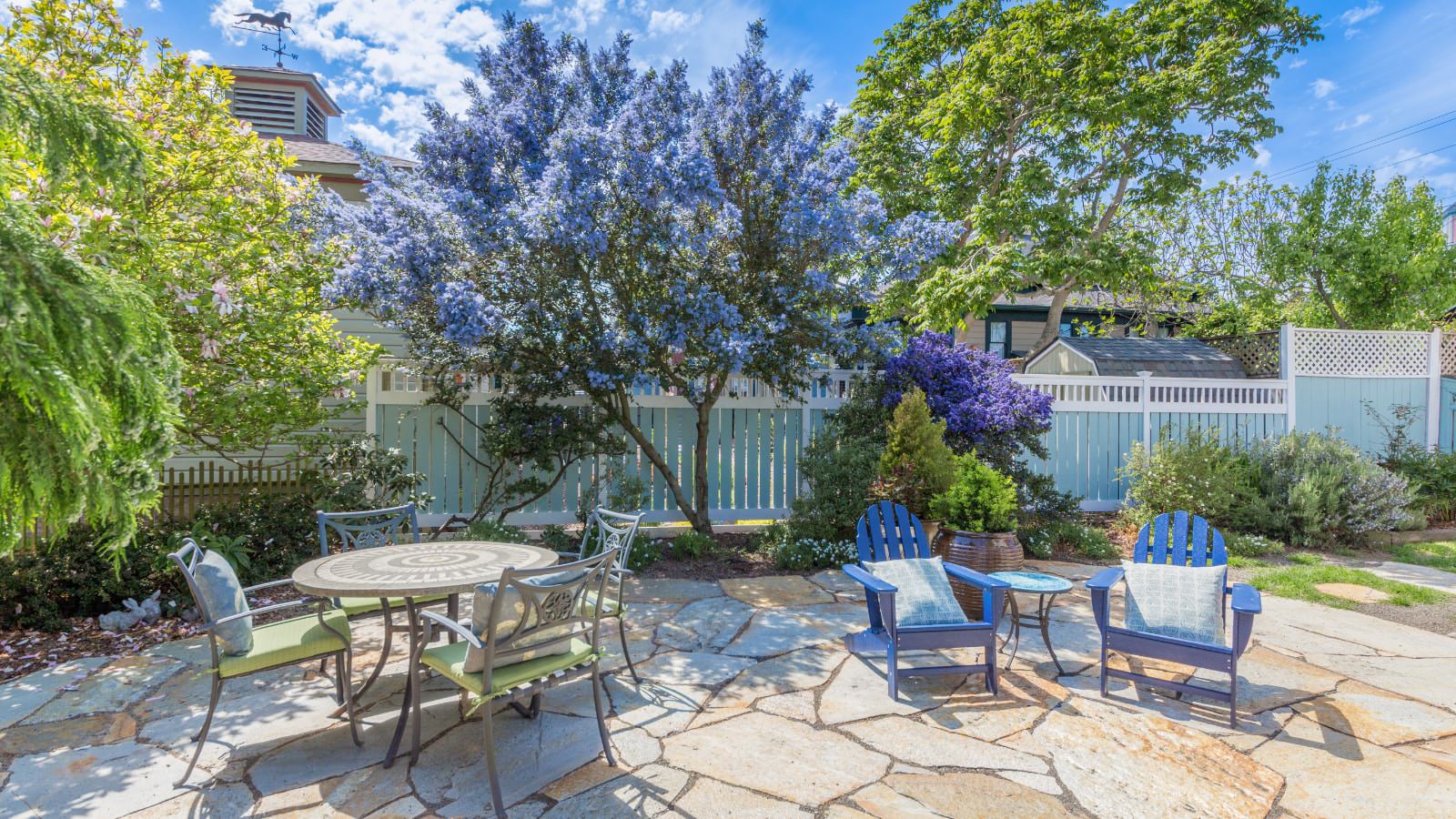 Stone patio with patio table and chairs, two blue Adirondack chairs, and surrounded by flower gardens and flowering bushes