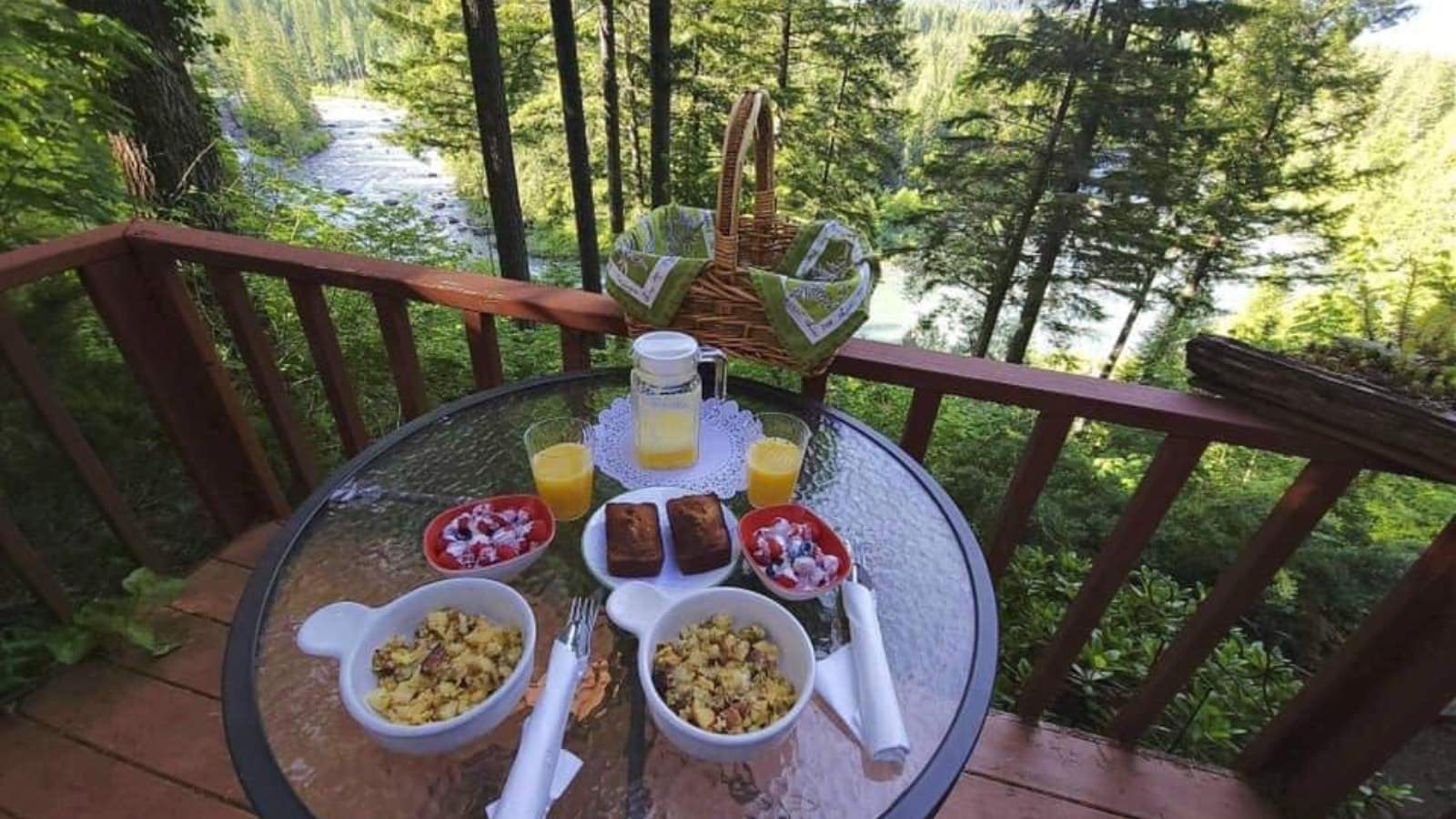 Red wooden deck with glass round patio table set for two looking out to a river surrounded by green bushes and trees