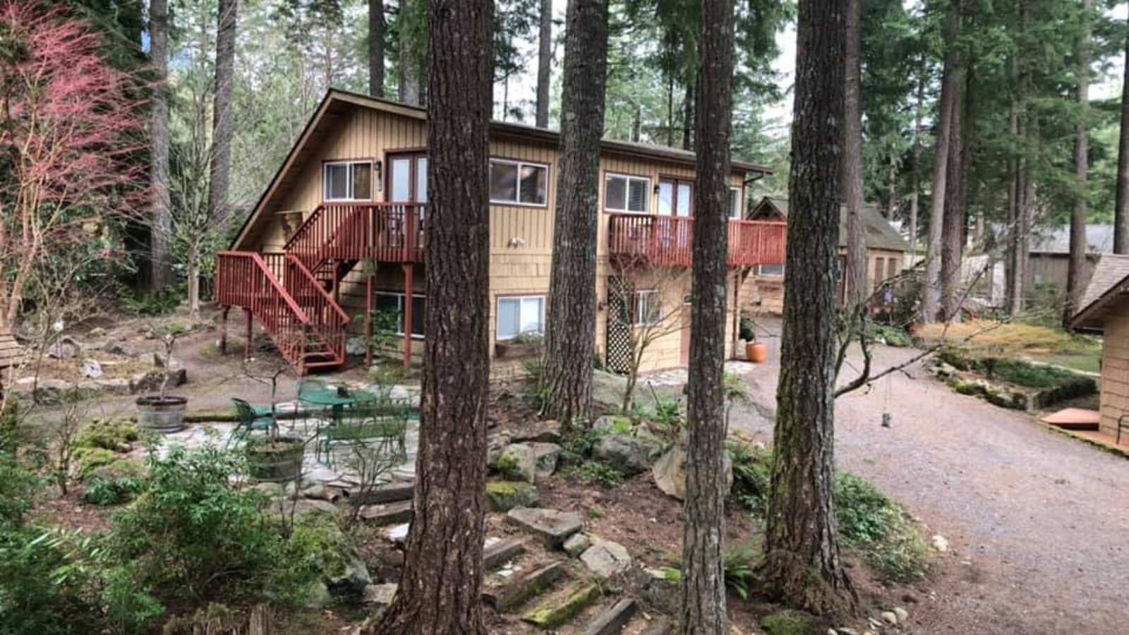 Exterior view of the property painted tan with brown trim, red deck with stairs, patio, and surrounded by large trees