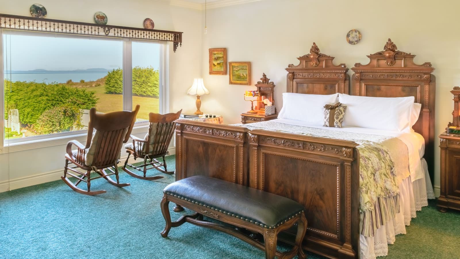 Bedroom with white walls, green carpeting, ornate wooden bed, light colored bedding, and two wooden rocking chairs looking out of large window