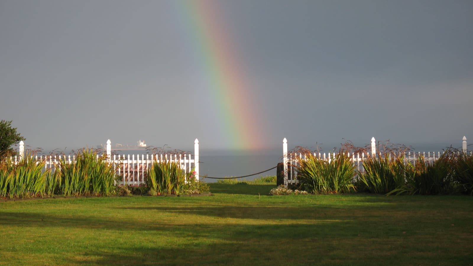 Backyard with green grass, white picket fence, view of the water, and rainbow in the background