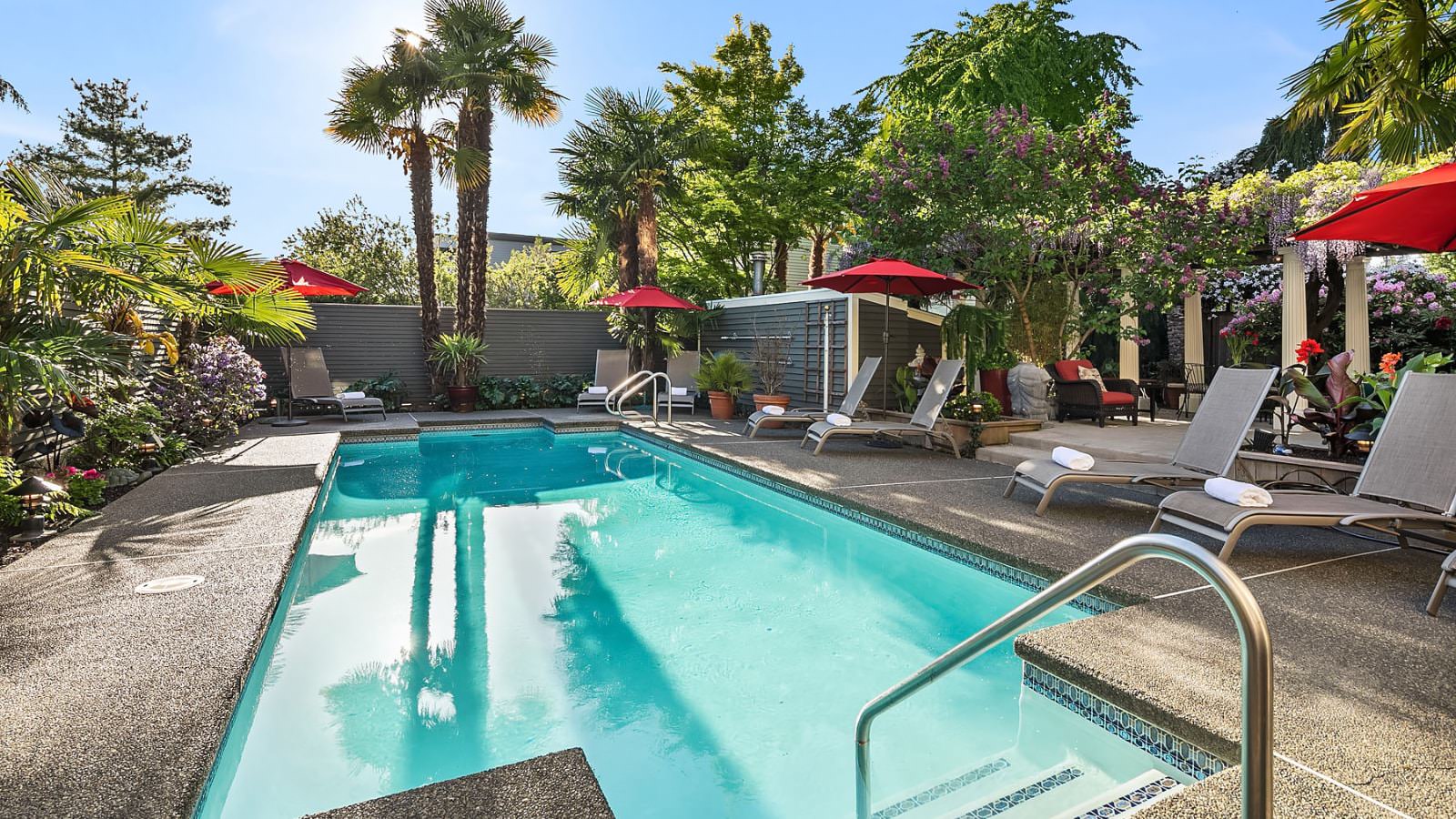 Pool with multiple lounge chairs, red umbrellas, bushes, flowers, and trees