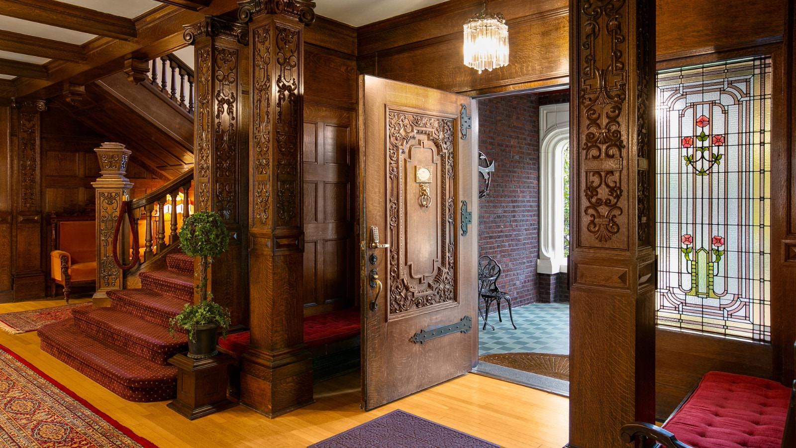 Front entrance of property with large ornate wooden door, huge ornate wooden staircase with red carpeted stairs, hardwood flooring, and dark wooden walls all around