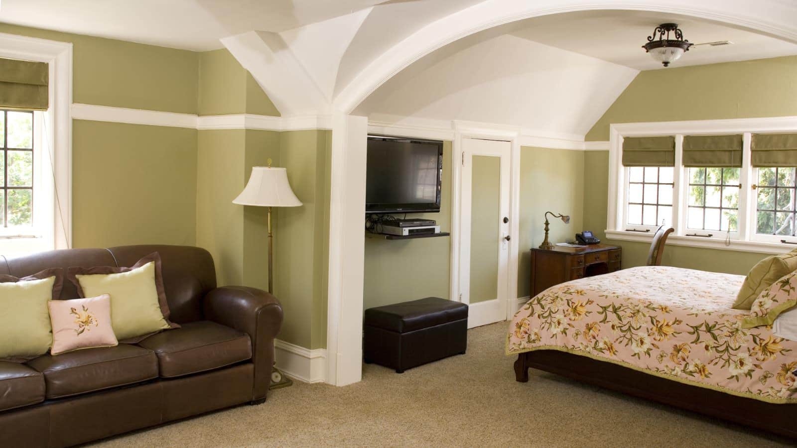 Bedroom suite with light green walls, white trim, light-colored carpeting, leather sofa, dark wooden bed, floral beddding, and wall-mounted flat-screen TV