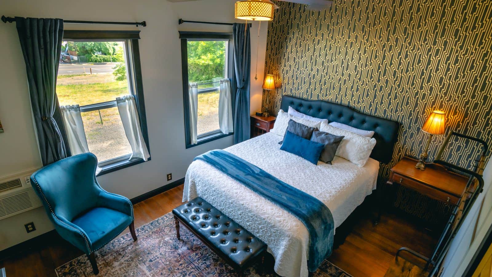 Bedroom with art deco black and gold wallpaper accent wall, hardwood flooring, black leather headboard, white bedding, blue upholstered antique armchair, and large windows