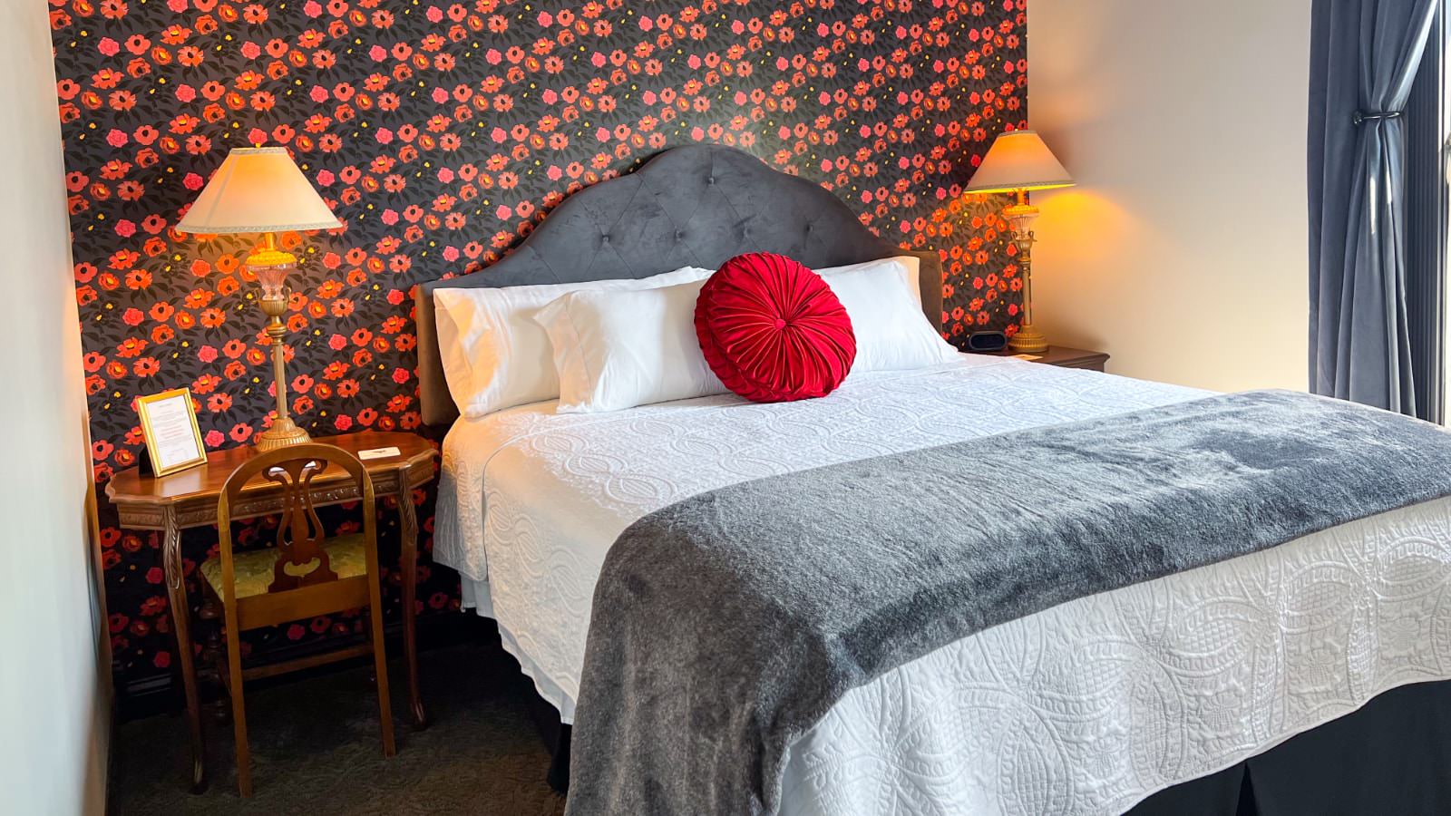 Bedroom with white walls, pink and black flowered wallpaper accent wall, upholstered head board, white bedding, and antique desk and chair as nighstand