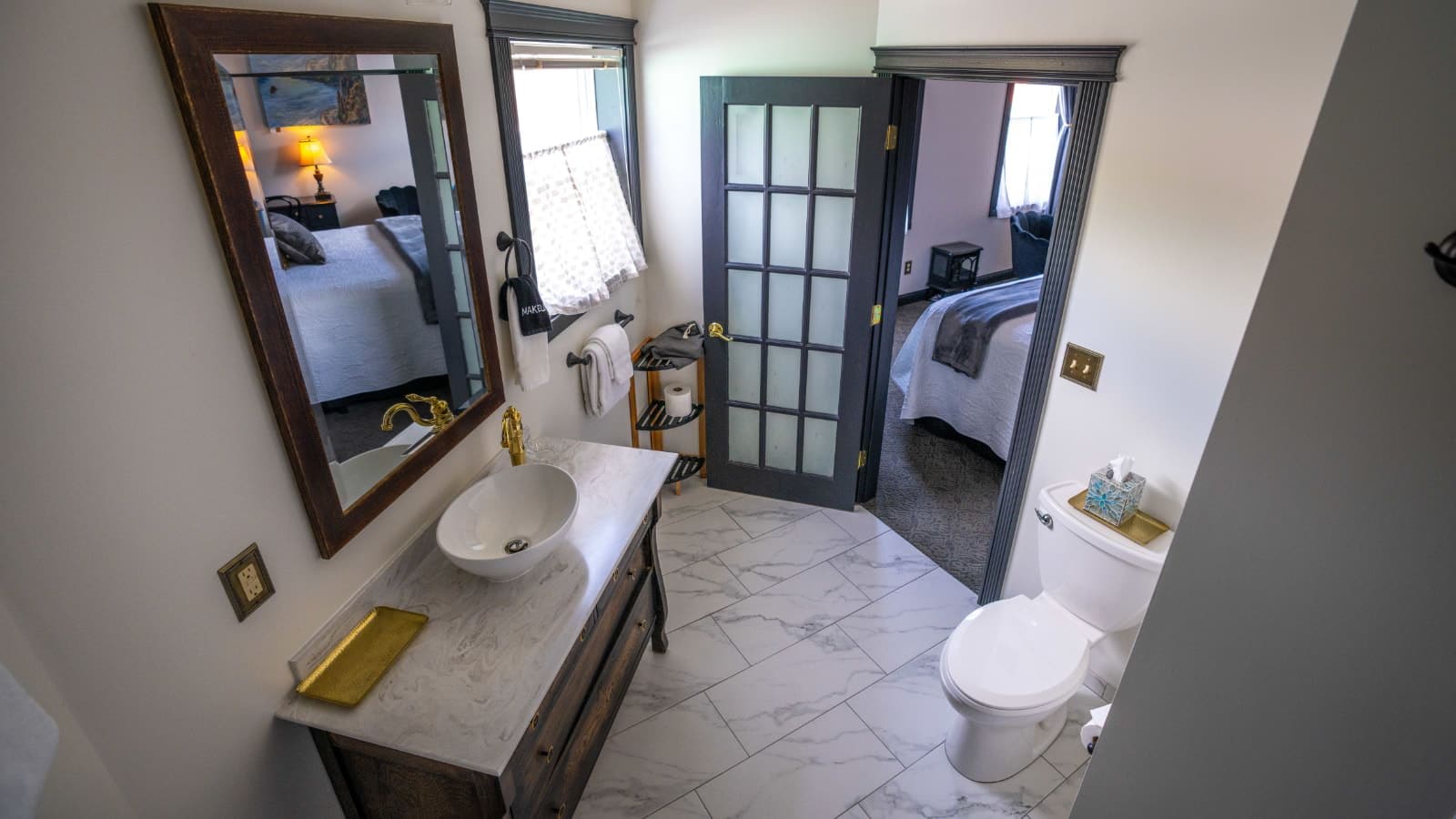 Bathroom with white walls, gray trim, marble tiled flooring, white toilet, wooden vanity, white bowl counter top sink, and wooden framed mirror