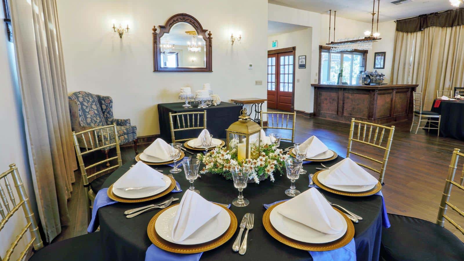 Dining area with white walls, hardwood flooring, round tables covered with black tablecloths, multiple place settings, and gold wooden chairs with black cushions