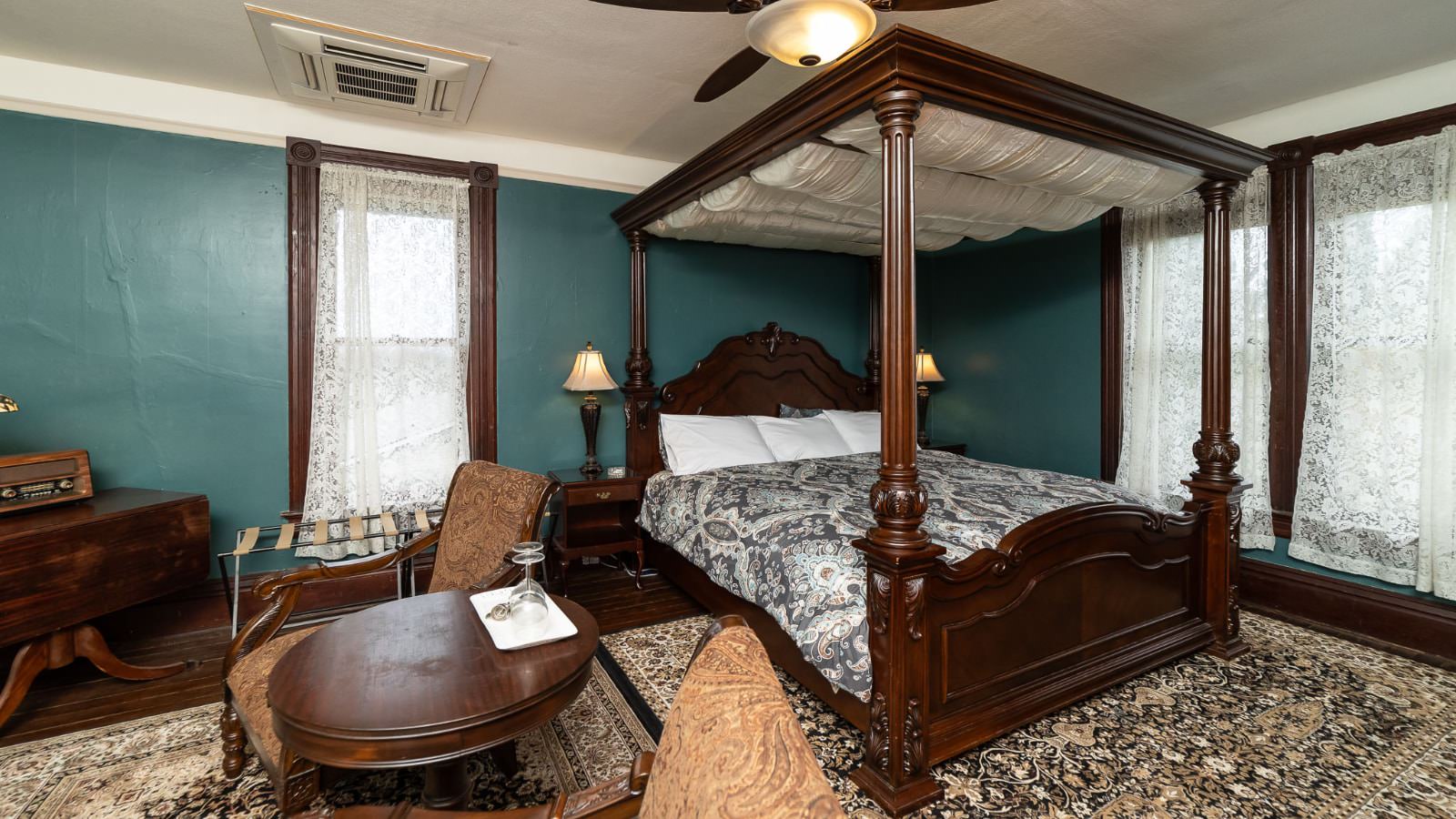 Bedroom with green walls, wood trim, hardwood flooring, dark wooden four-poster bed with canopy, multicolored bedding, sitting area, and large windows