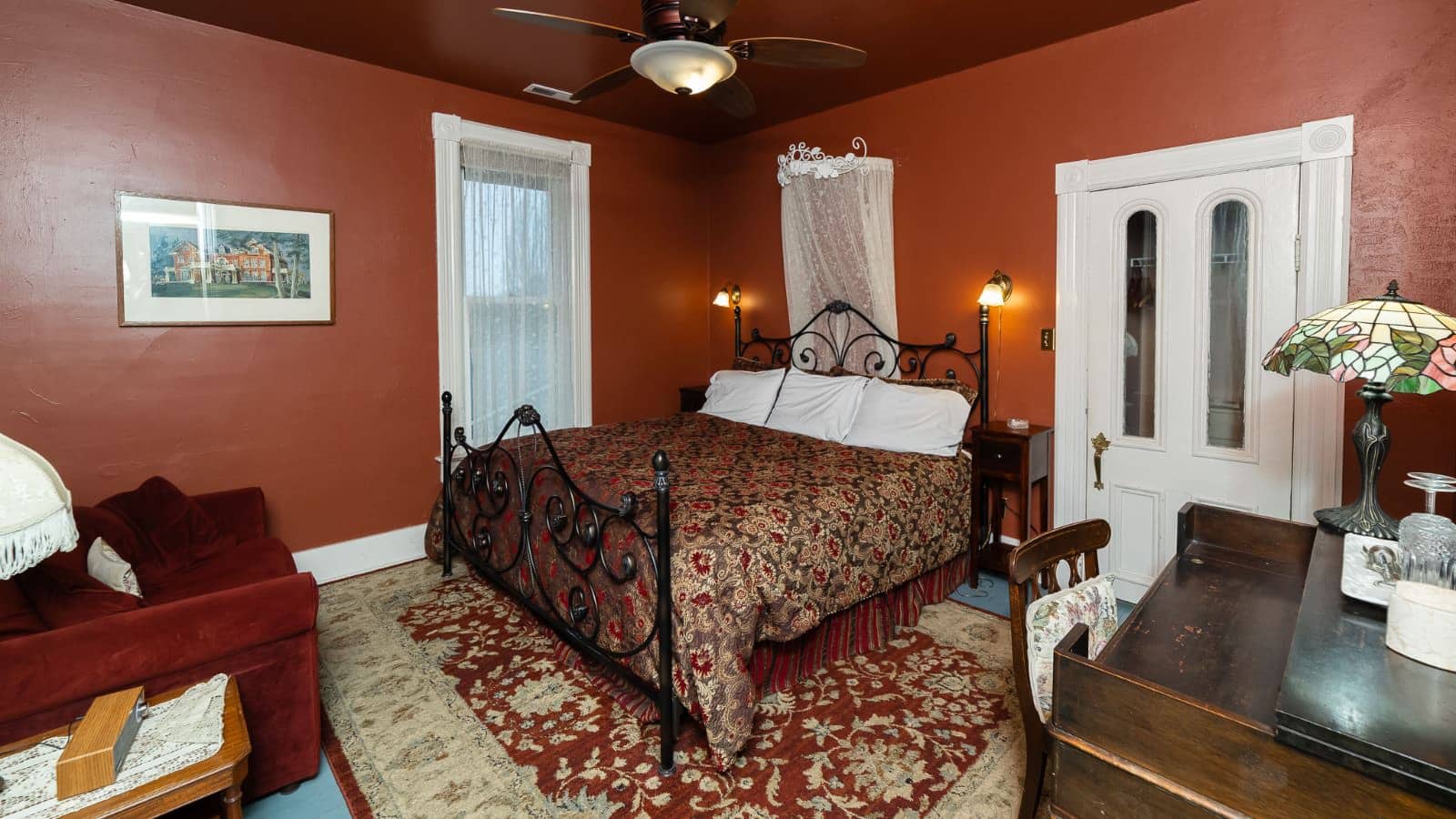 Bedroom with rust walls, hardwood flooring, wrought iron bed, multicolored bedding, antique wooden desk, and red velvet upholstered chair