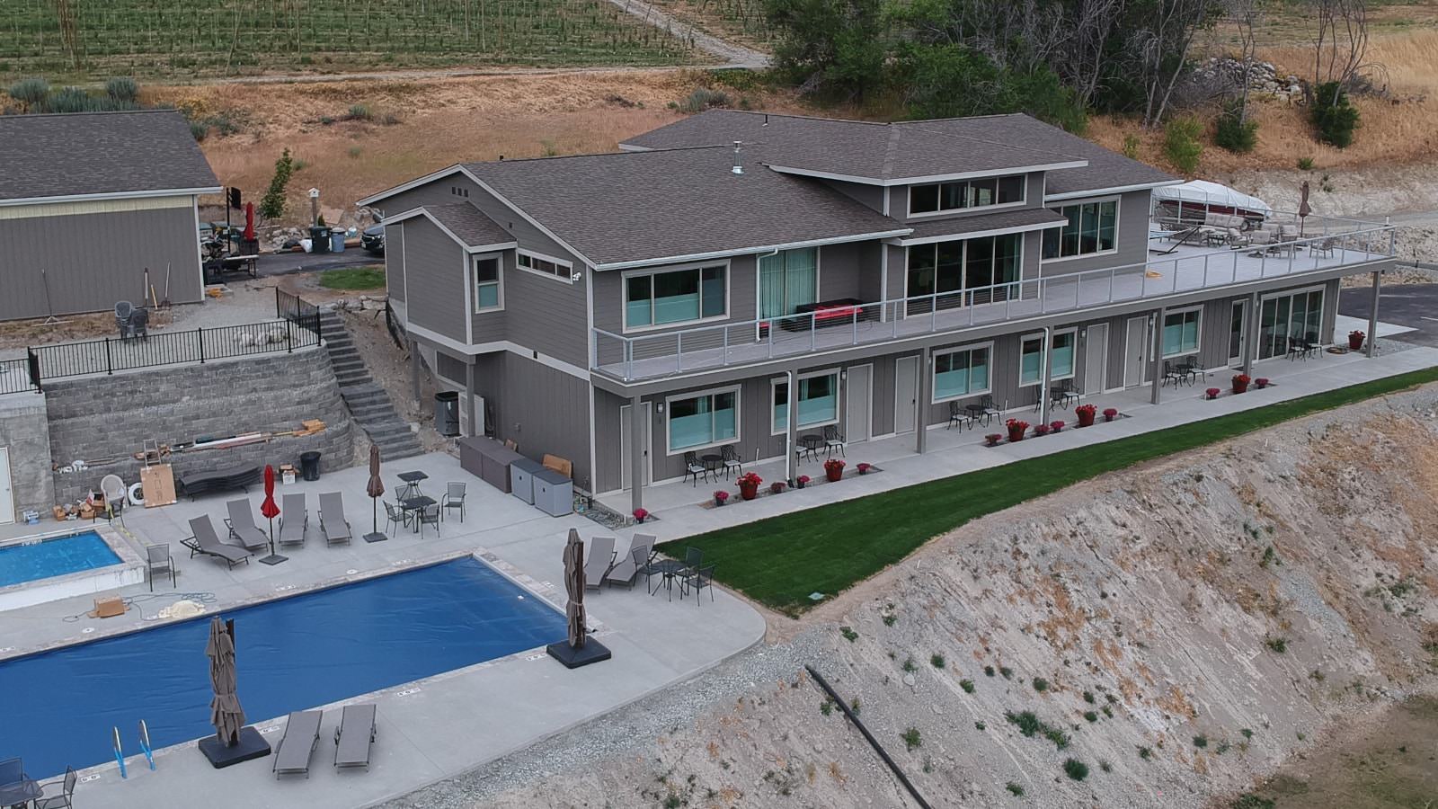 Aerial view of property painted dark gray with white trim, second level wrap-around deck, green grass, pool, and hot tub all sitting on the edge of a rocky hill