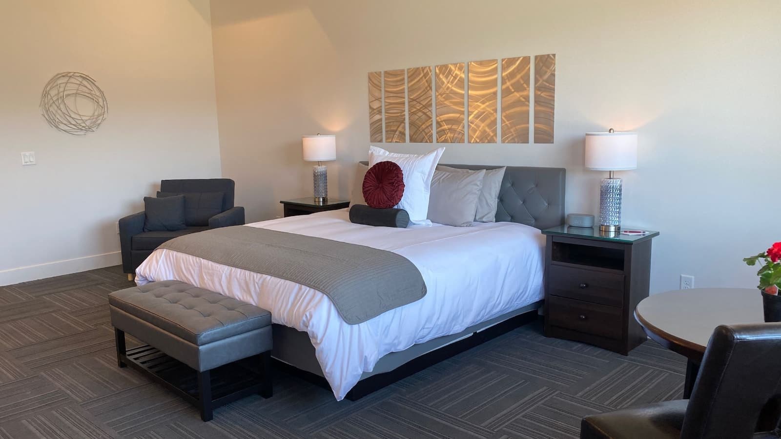 Bedroom with white walls, gray carpeting, gray leather headboard, white bedding, gray upholstered armchair, and dark wooden nightstands with lamps