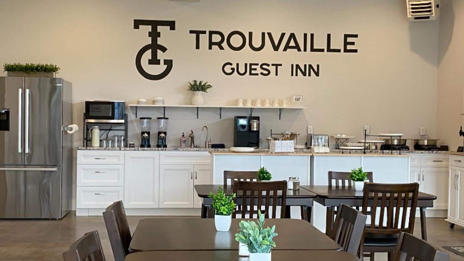 Large dining area with cream walls, concrete flooring, white cabinets, large counter top, stainless steel refrigerator, dark wooden tables and chairs, and Trouvaille Guest Inn text in black on wall