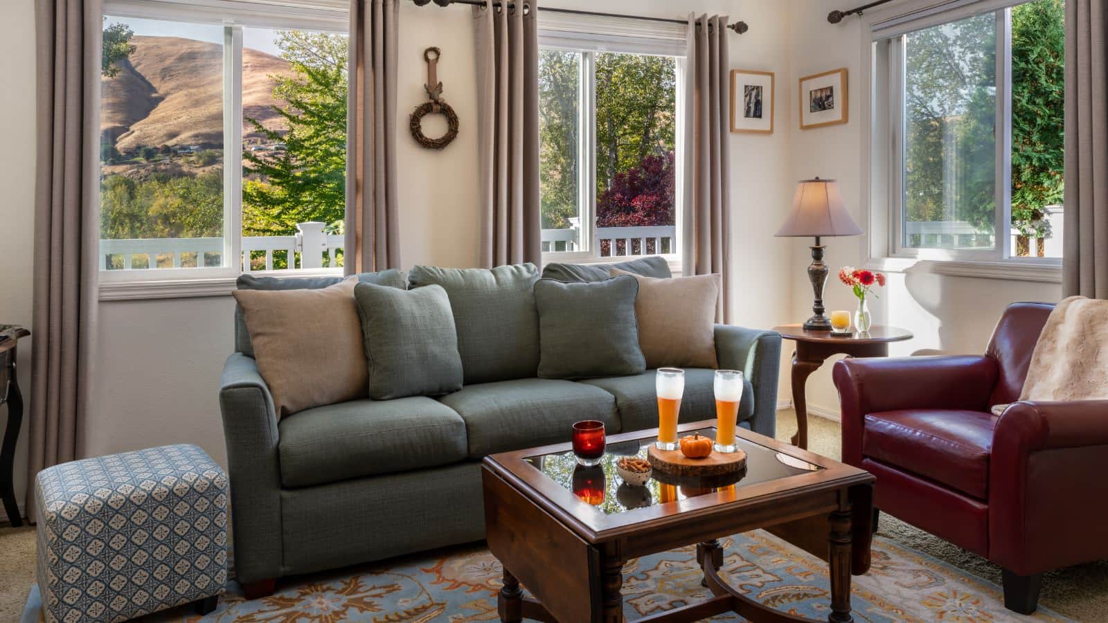 Living room with white walls, white trim, carpeting, sage green upholstered sofa, red leather armchair, and large windows with views of the hills in the background