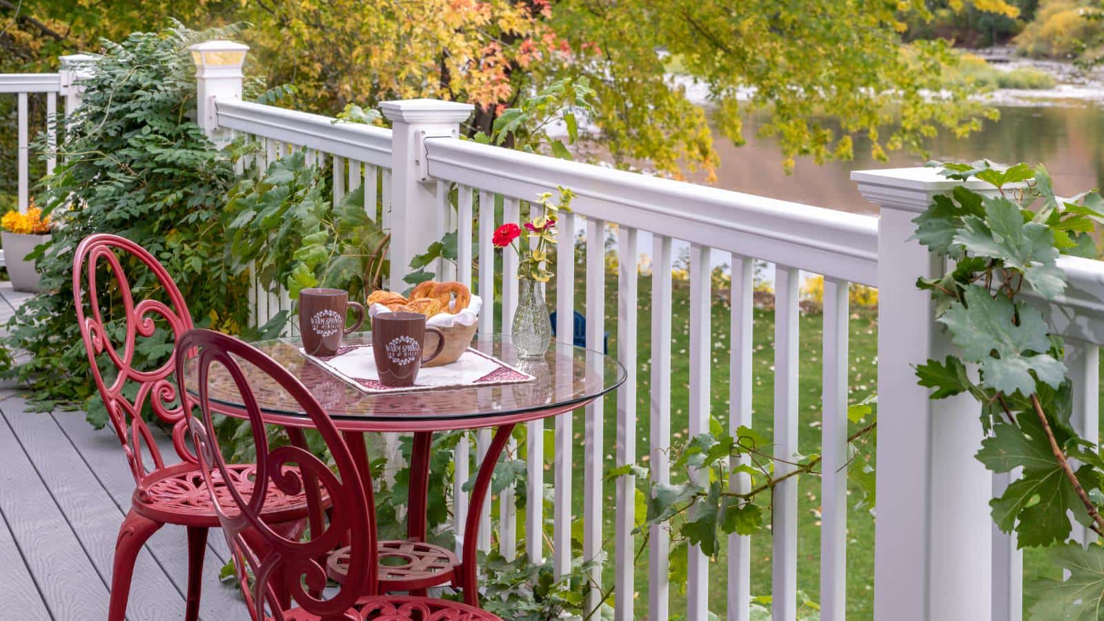 Close up view of red wrought iron patio table and chairs on deck with white railing looking out to the grass and water below