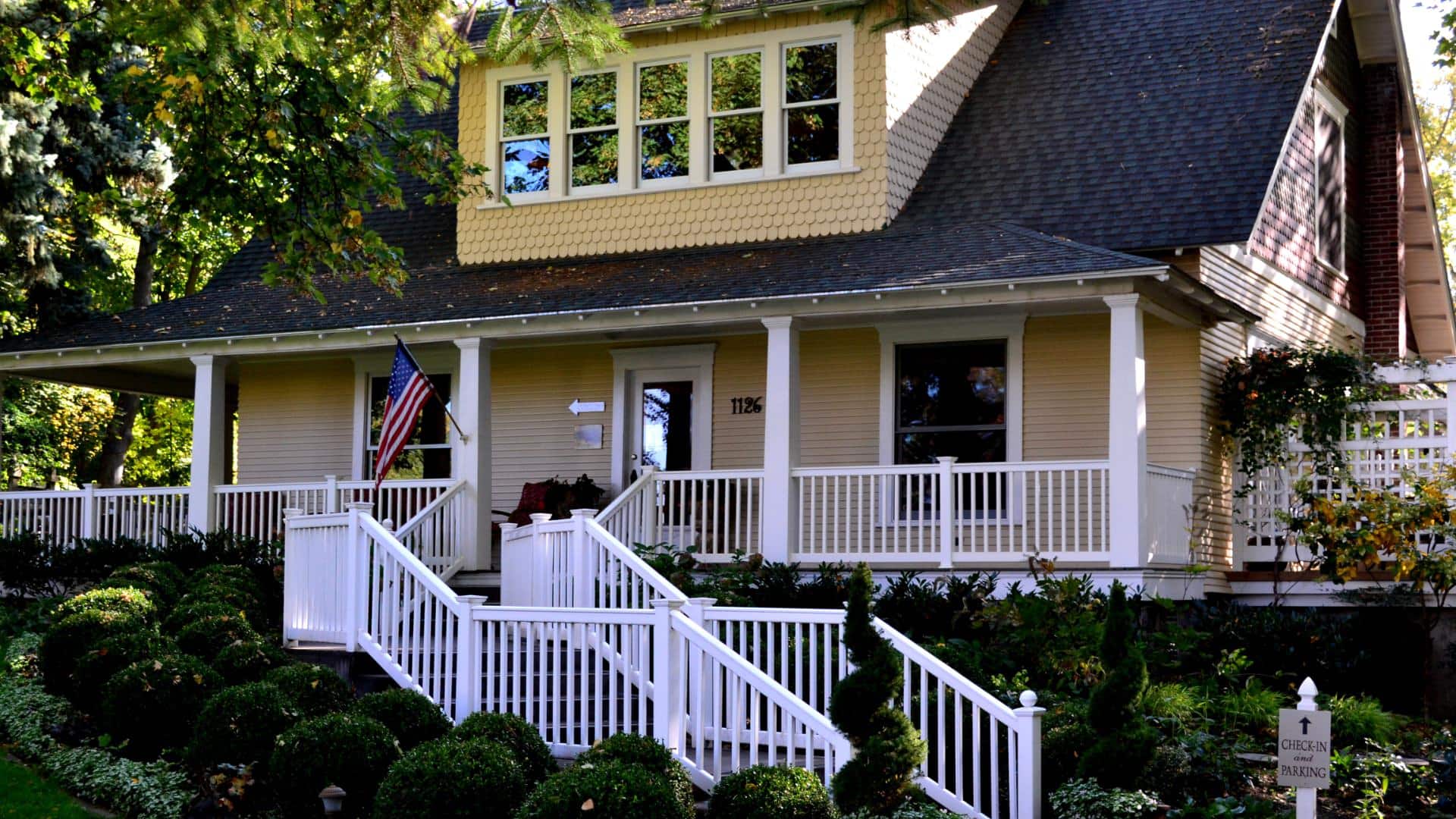 Exterior view of property painted cream with white trim, wrap-around porch, white railing, stairs leading to front entry, and surrounded by green bushes and trees