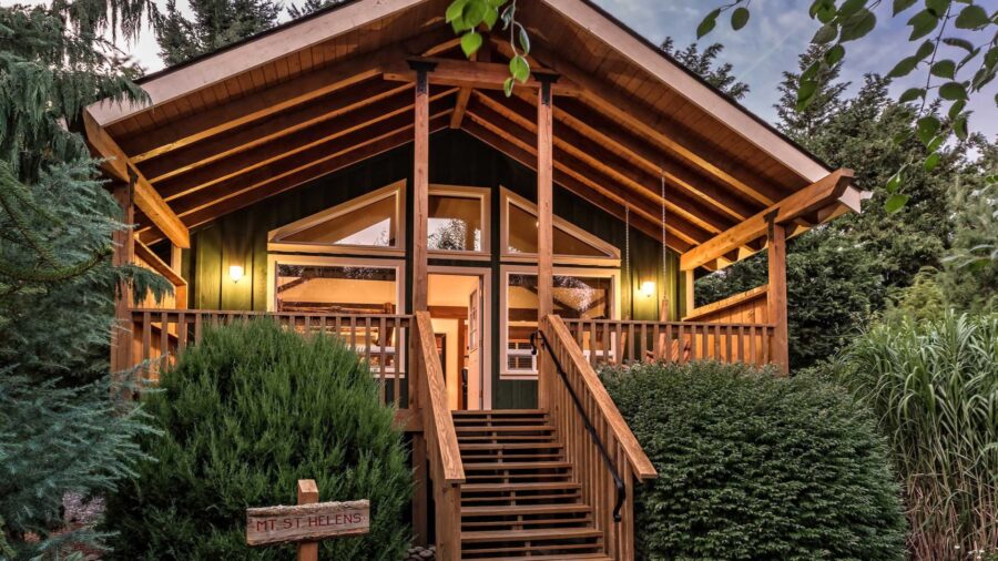 Exterior view of property painted green with natural wood trim and railings surrounded by green bushes, grasses, and trees