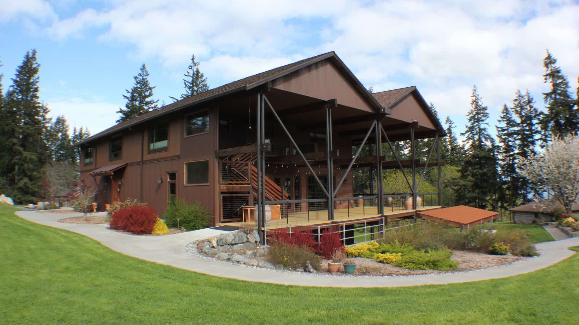 Exterior view of property painted brown with large deck surrounded by green grass and multicolored bushes