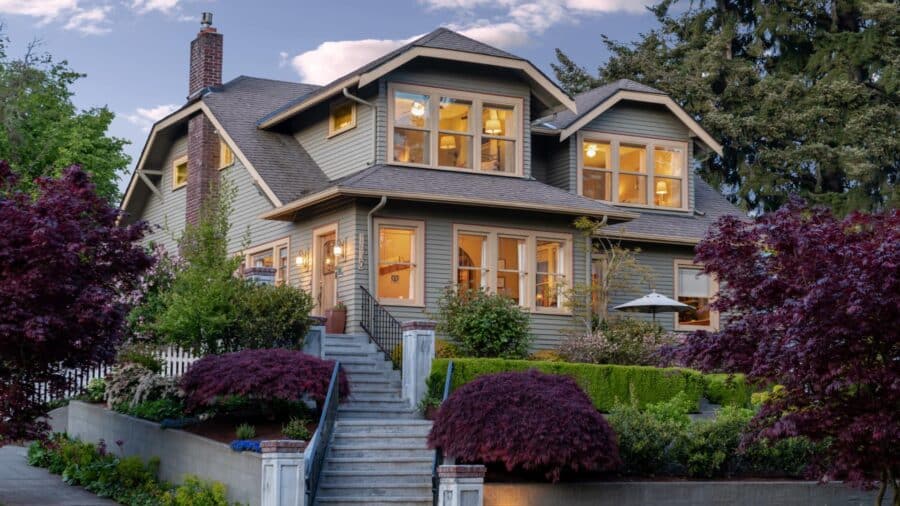 Exterior view of property with light gray siding, almond trim, stairs up to entry and surrounded by lush gardens, bushes, and trees