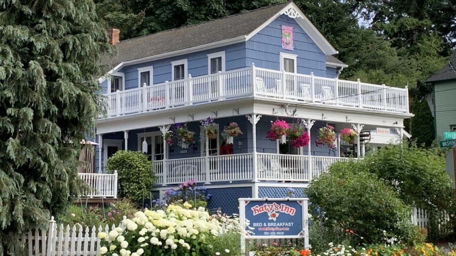 Exterior view of property painted light blue with white trim, white railed front porch and second-level balcony, and surrounded by green bushes, flowers, and trees