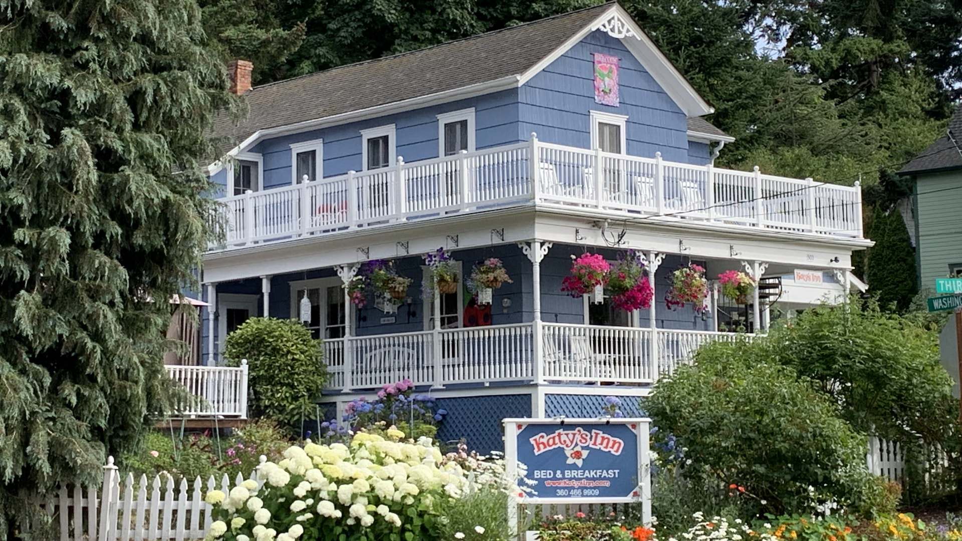 Exterior view of property painted light blue with white trim, white railed front porch and second-level balcony, and surrounded by green bushes, flowers, and trees