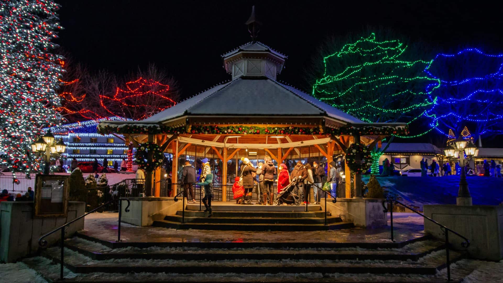 People standing in a lighted gazebo at night looking at homes and trees decorated with Christmas lights