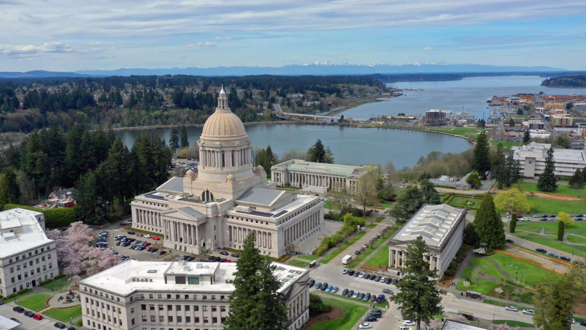 Aerial view of a Capitol building surrounded by other buildings, green trees, bodies of water, and mountain range in the background