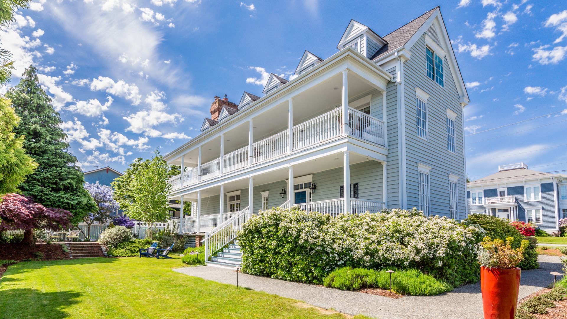 Exterior view of property painted light blue with white trim, white front porch, second-level balcony and surrounded by green grass, lush bushes, and trees